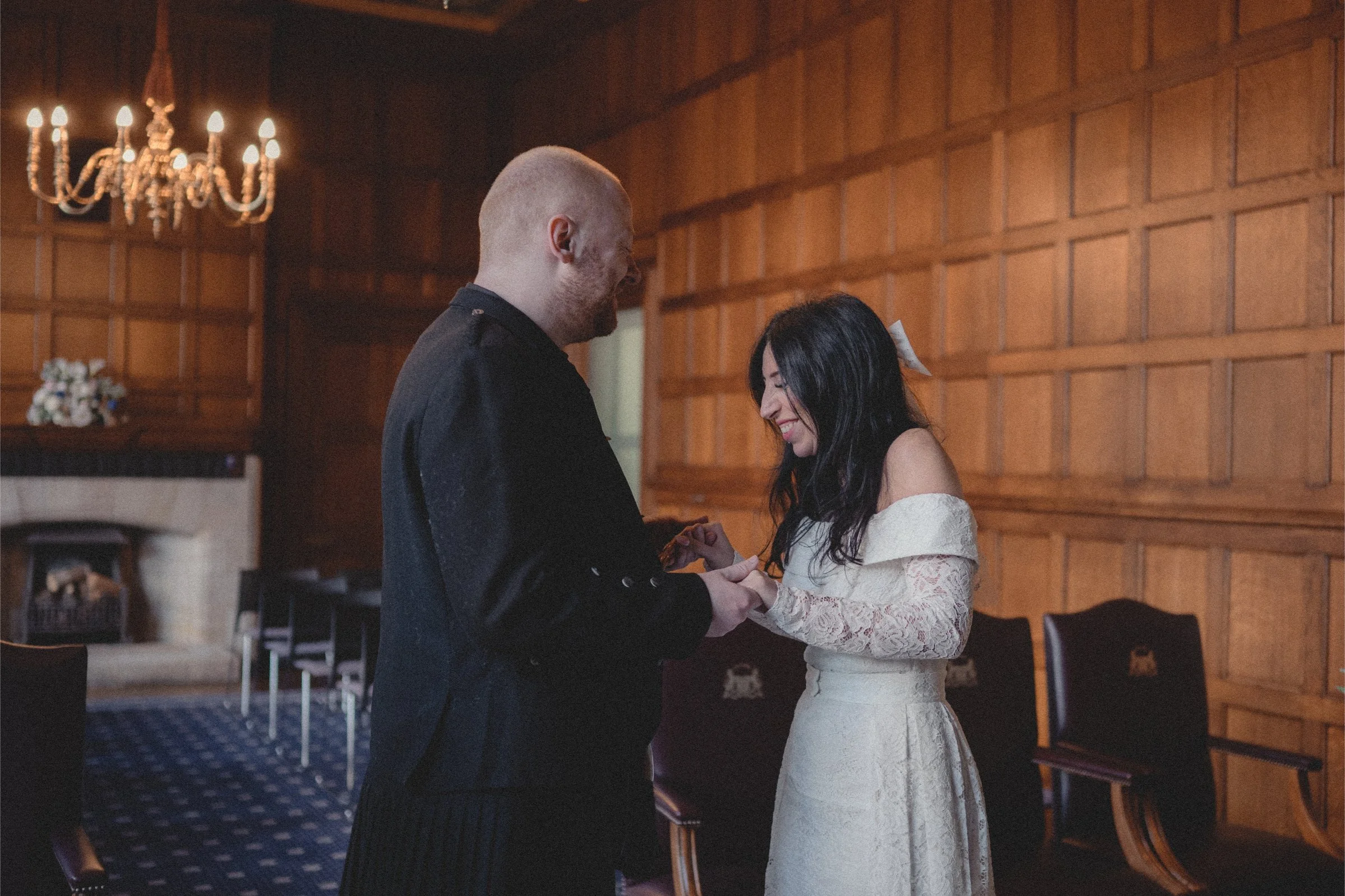 A couple holding hands on their wedding day indoors with wood-paneled walls and a chandelier in the background.