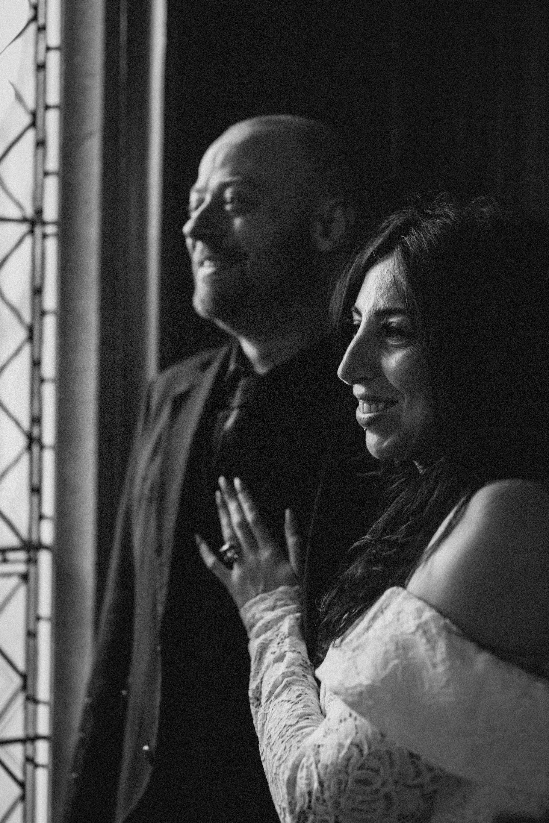 A black and white photo of a smiling man and woman standing side by side, with the woman touching the man's chest, both looking to the right.