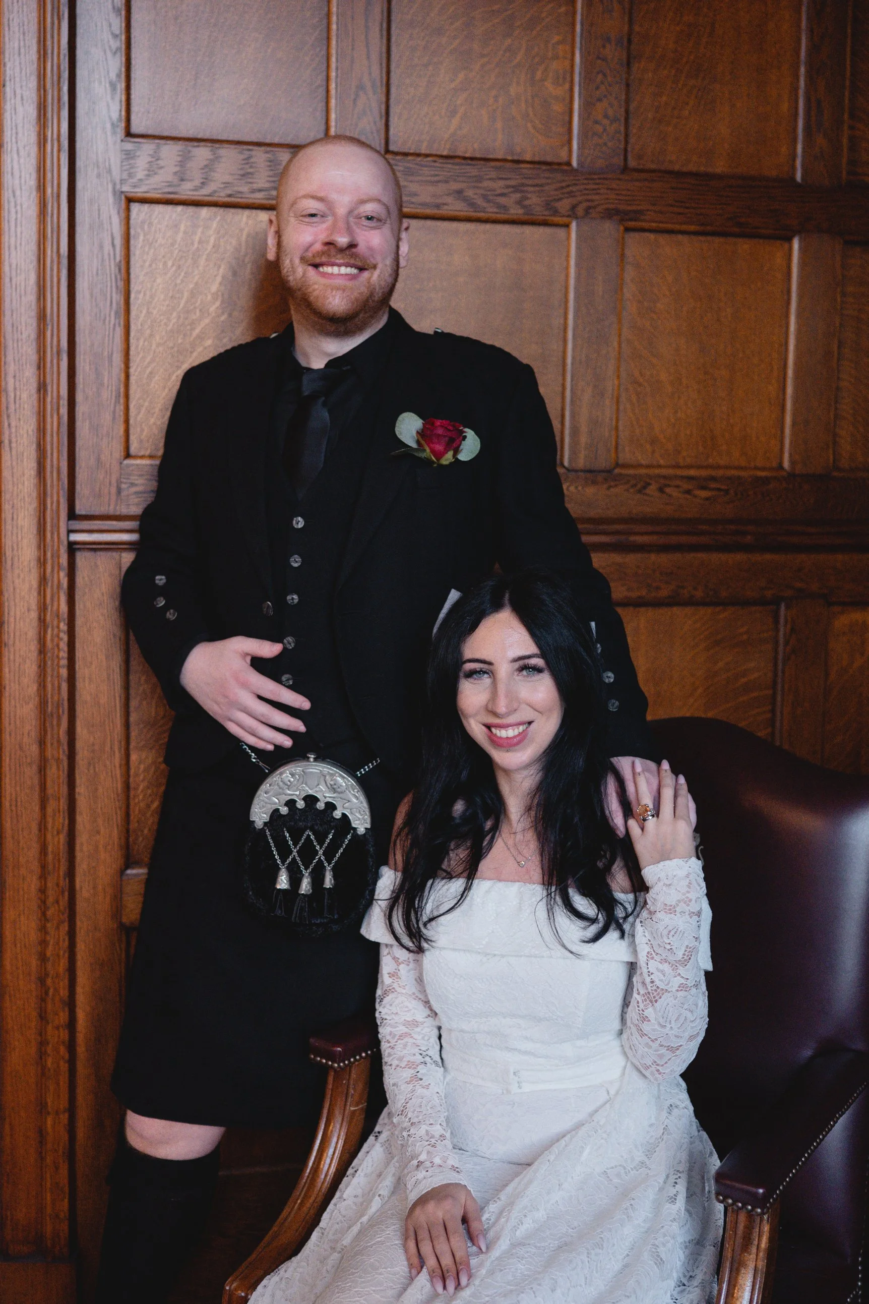 A man in a black suit and tie with a red rose boutonniere, standing behind a woman in a white lace dress, sitting in a wooden chair in front of a wooden-paneled wall.
