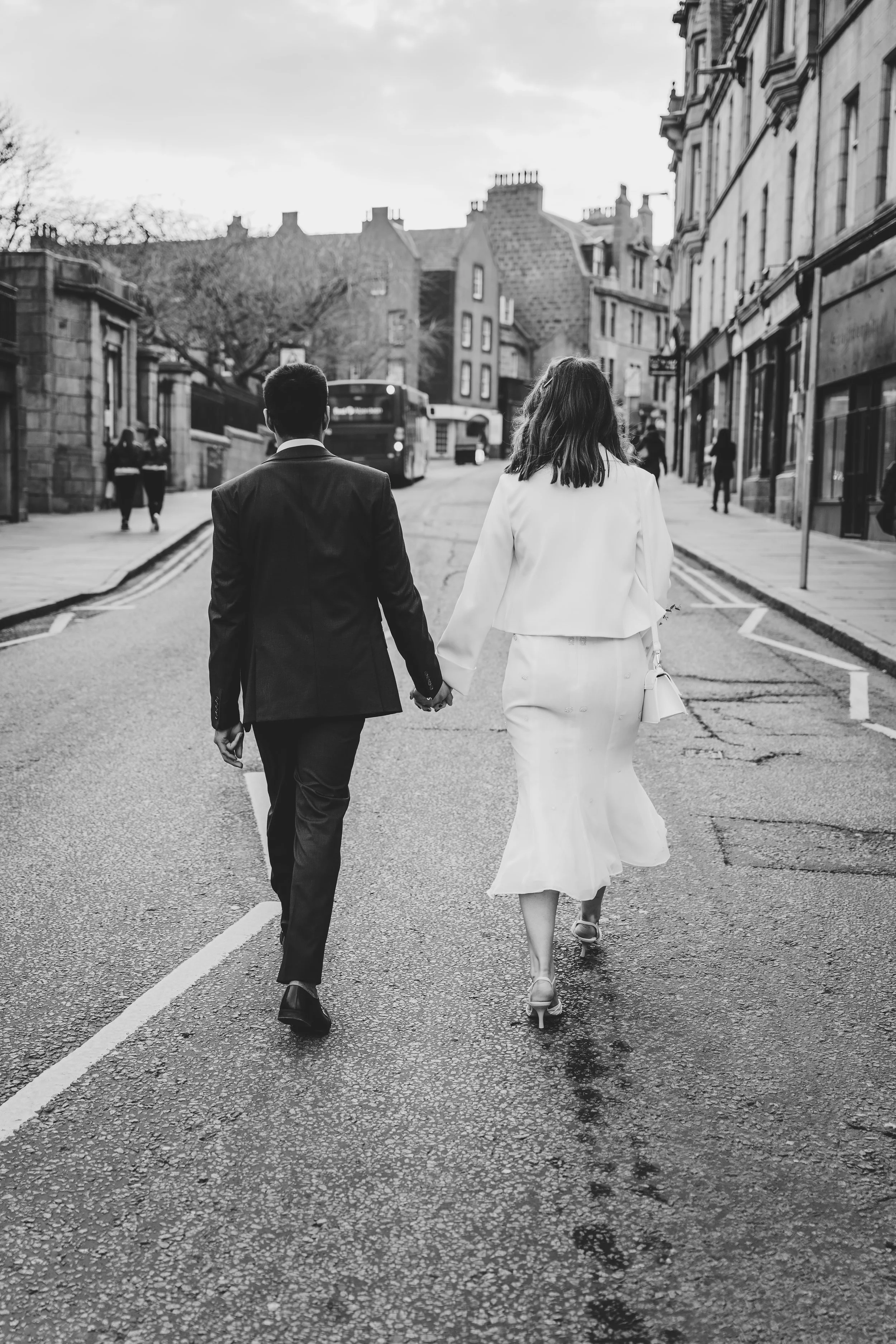 A black and white photo of a couple walking hand in hand on a city street, dressed in formal attire, with buildings and pedestrians in the background.