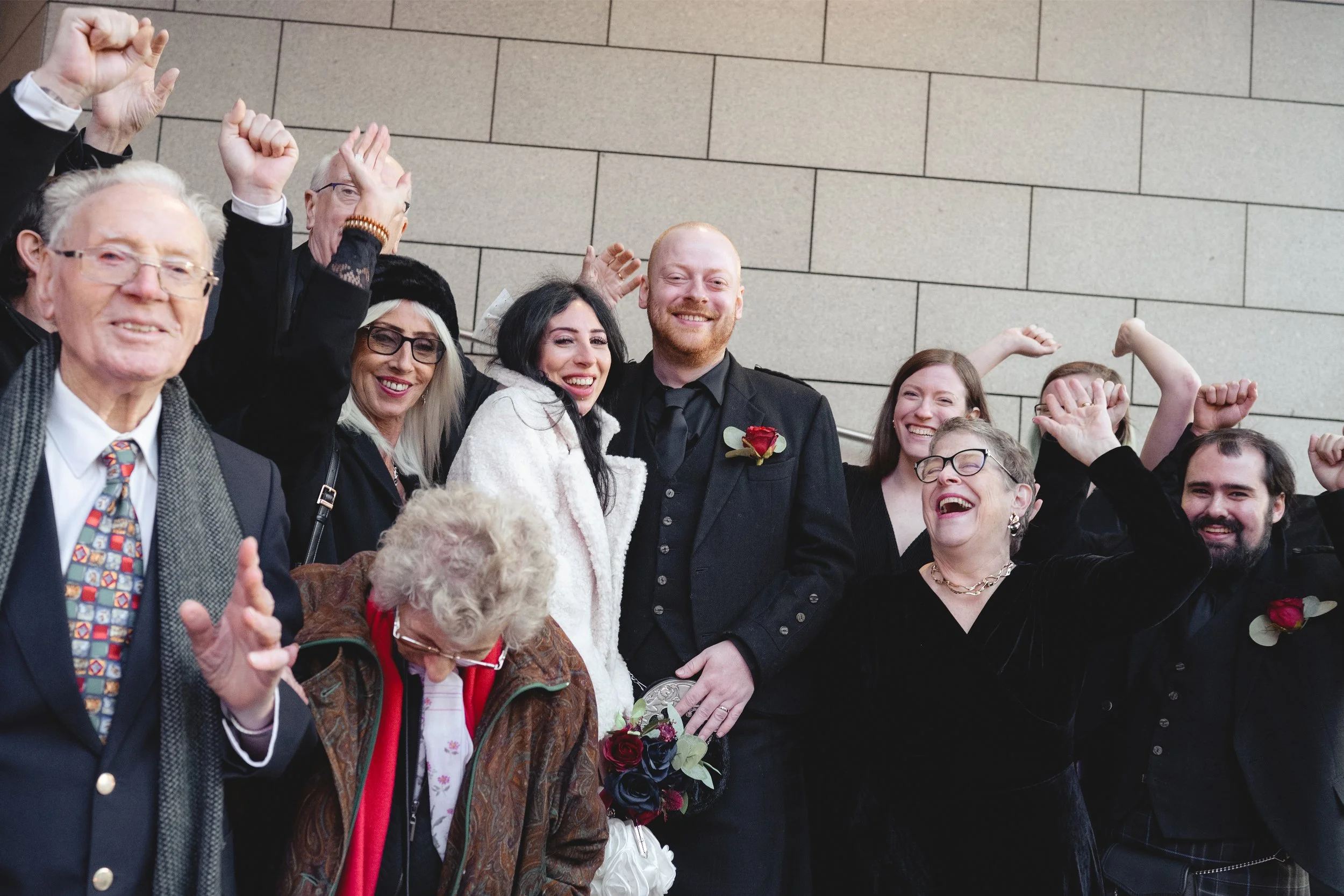 A group of people celebrating a wedding, with some raising their fists and smiling, standing against a beige tiled wall.