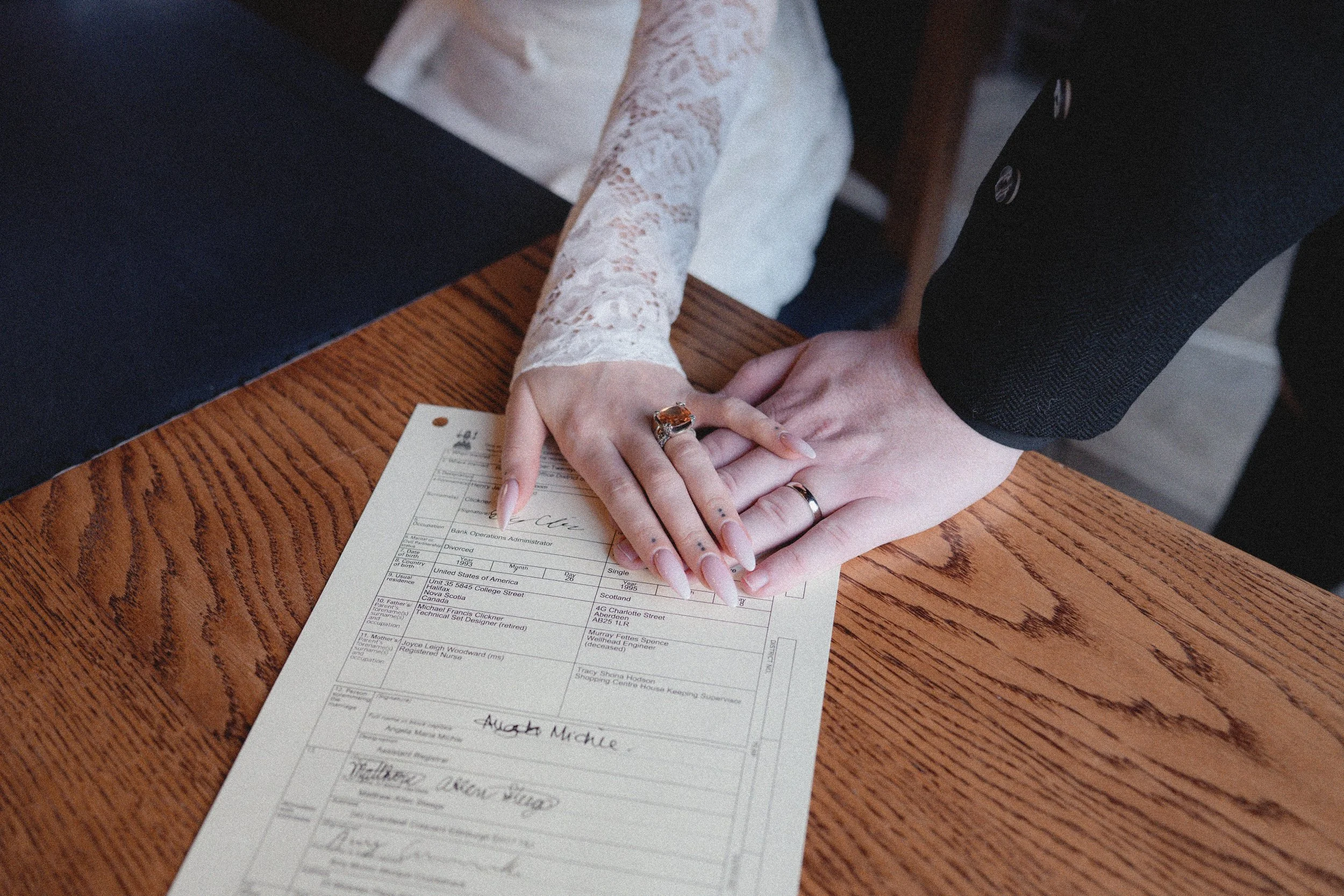 Couple's hands with wedding rings resting on a legal document on a wooden table, with the woman wearing a lace dress and the man in a black suit.