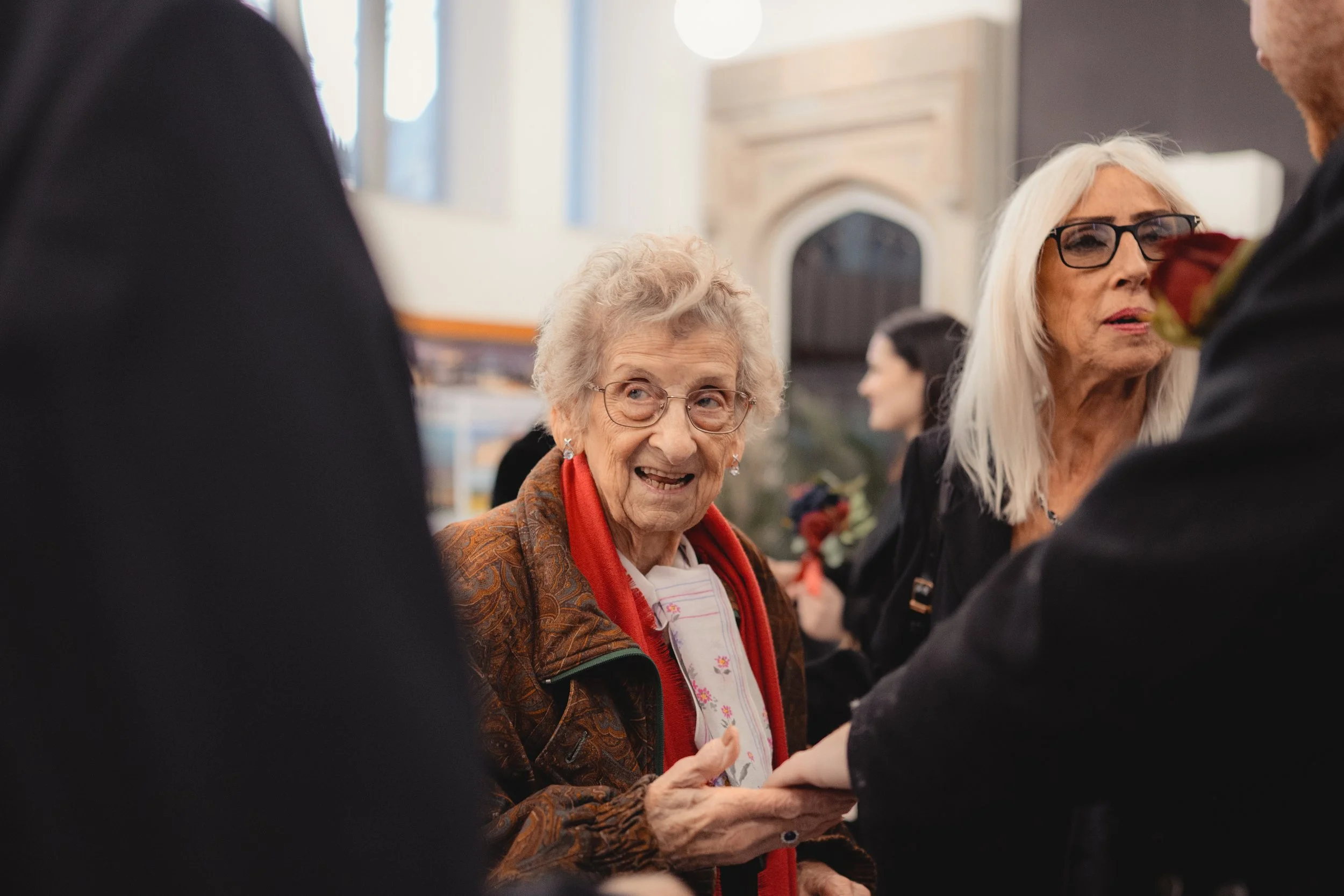 An elderly woman with glasses, curly gray hair, and a warm smile interacts with a man holding a rose in a social gathering indoors. Two other women are visible in the background, one with silver hair and glasses, and the other with dark hair, both en