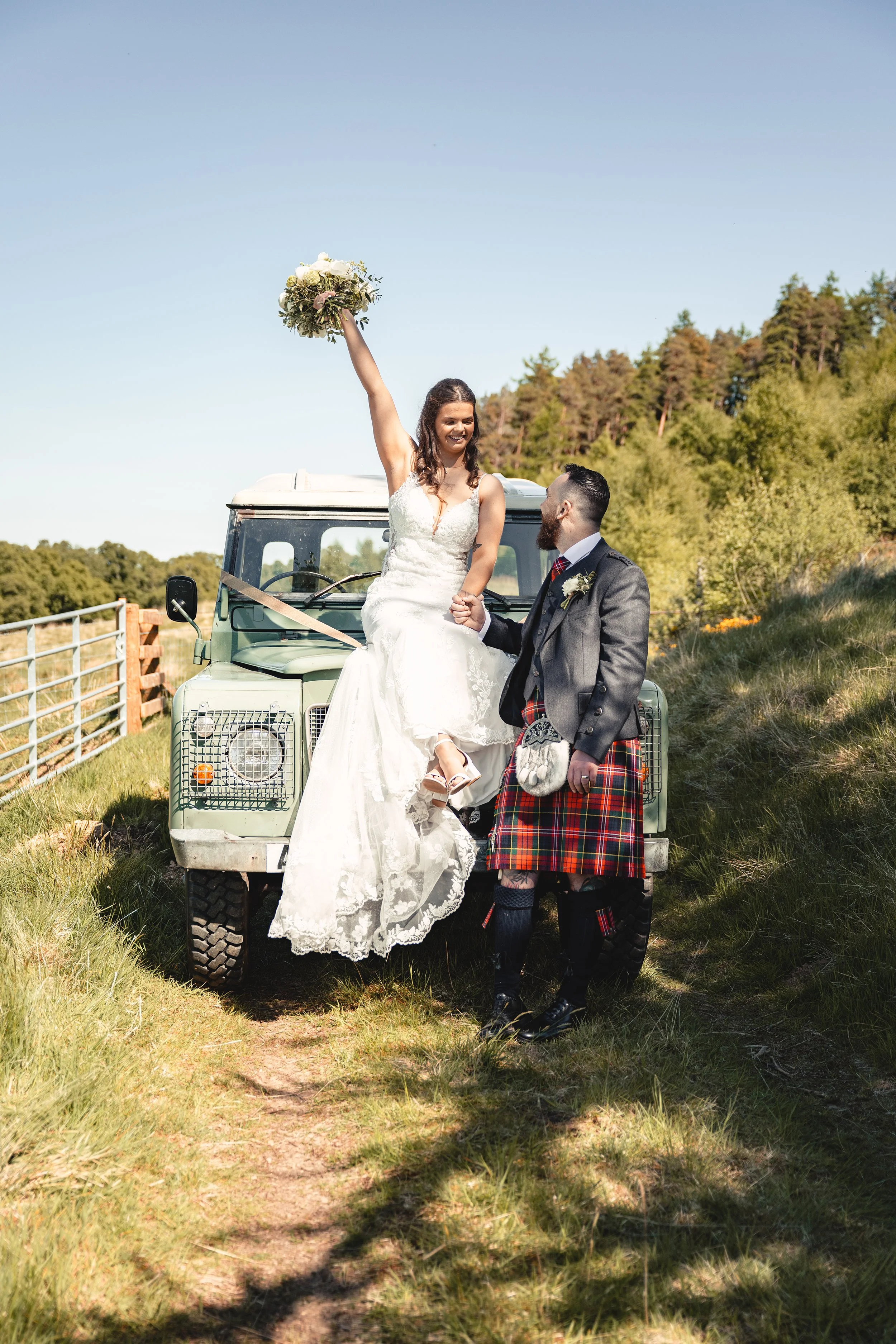 A bride in a white wedding dress sitting on the hood of a vintage green Land Rover, holding a bouquet of flowers high above her head, while a groom dressed in traditional Scottish attire, including a kilt, stands nearby holding her hand in a scenic o