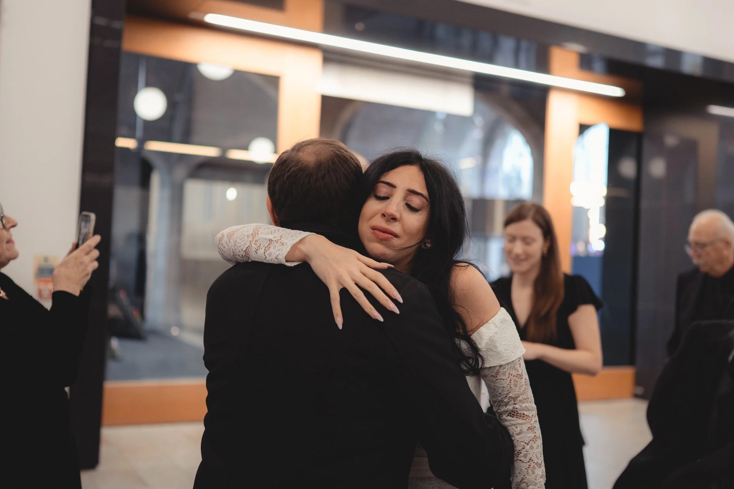 Woman with dark hair embraces man at a social gathering, expressing emotion; other guests are visible in the background, some smiling and one taking a photo.