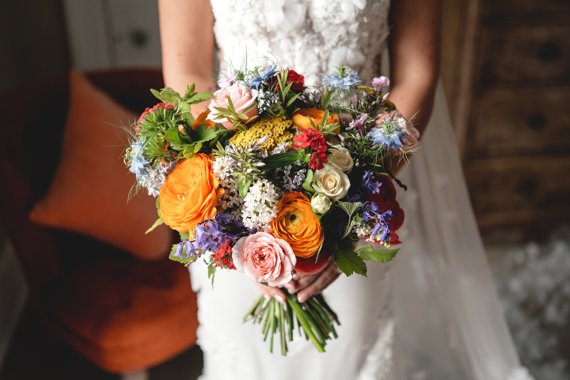 A bride holding a colorful bouquet of flowers, including roses, ranunculus, and various wildflowers, against a rustic indoor background.