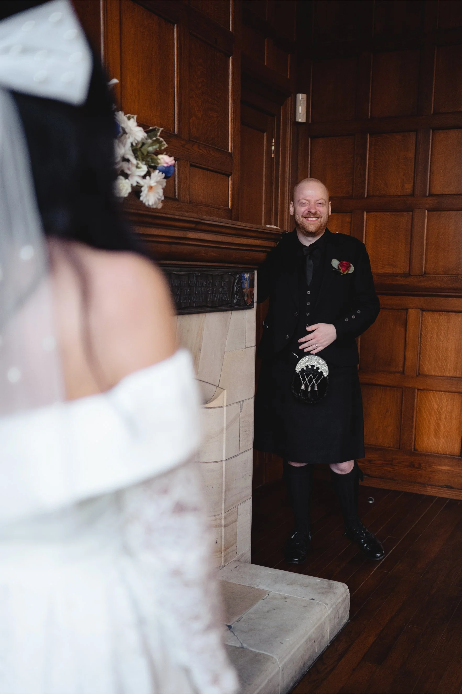 A man dressed in traditional Scottish attire, including a kilt, standing by a fireplace and smiling at a woman partially visible in the foreground, inside a wood-paneled room.