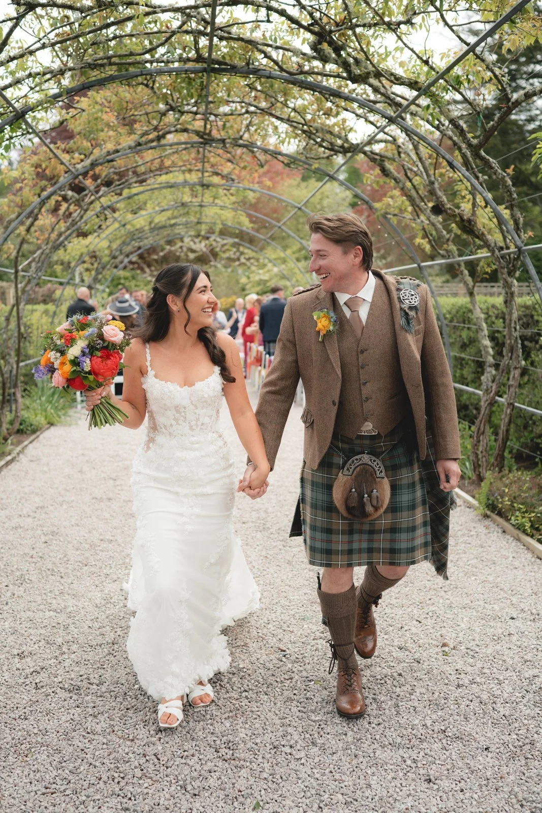 A bride and groom walking hand in hand outdoors, smiling at each other, under a vine-covered archway during a wedding celebration.