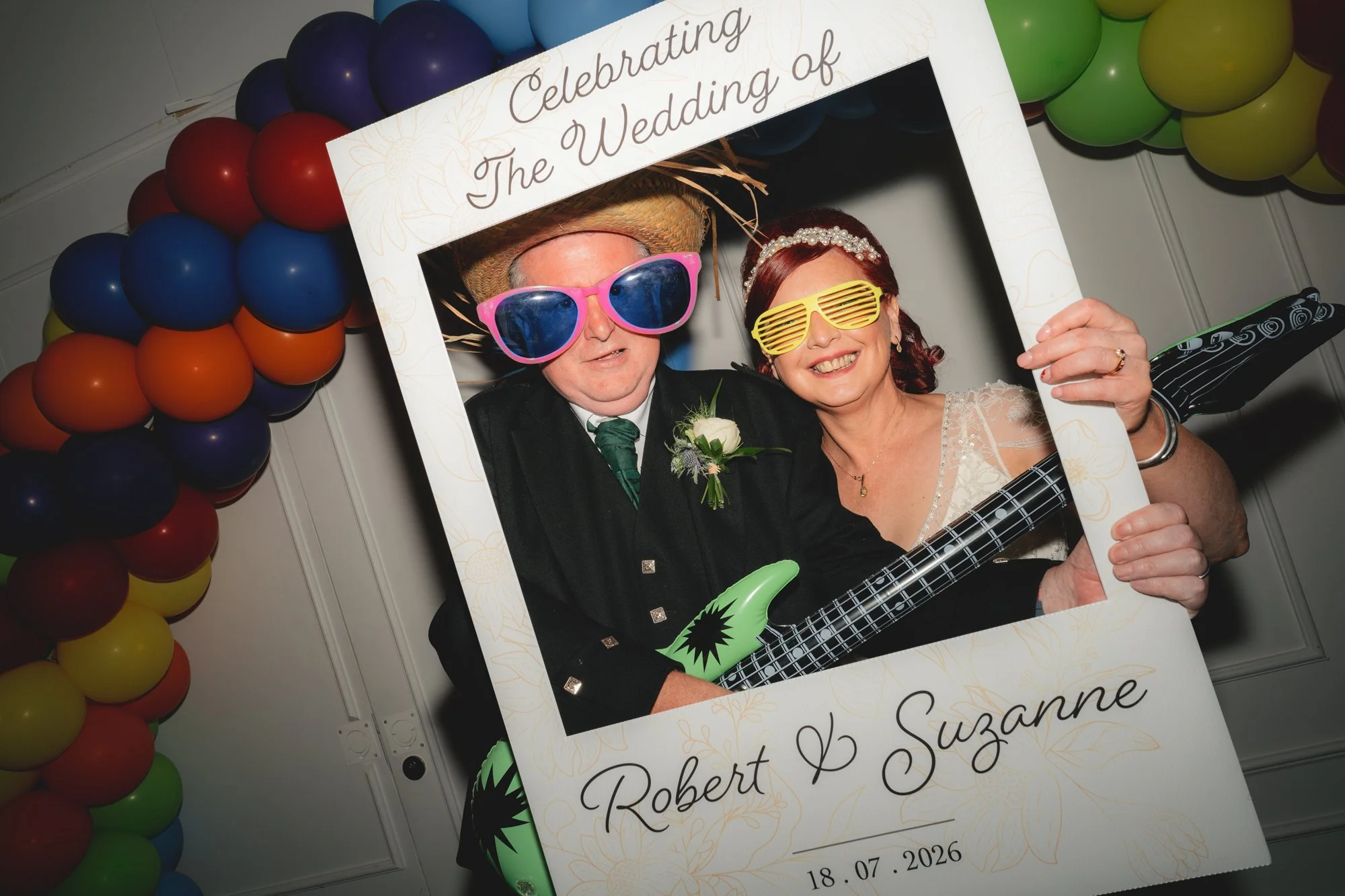 A couple at a wedding celebration holding a decorative frame with a sign that reads "Celebrating the Wedding of Robert & Suzanne" and the date "18.07.2026." The man is wearing oversized pink sunglasses and a straw hat, while the woman has on yellow s