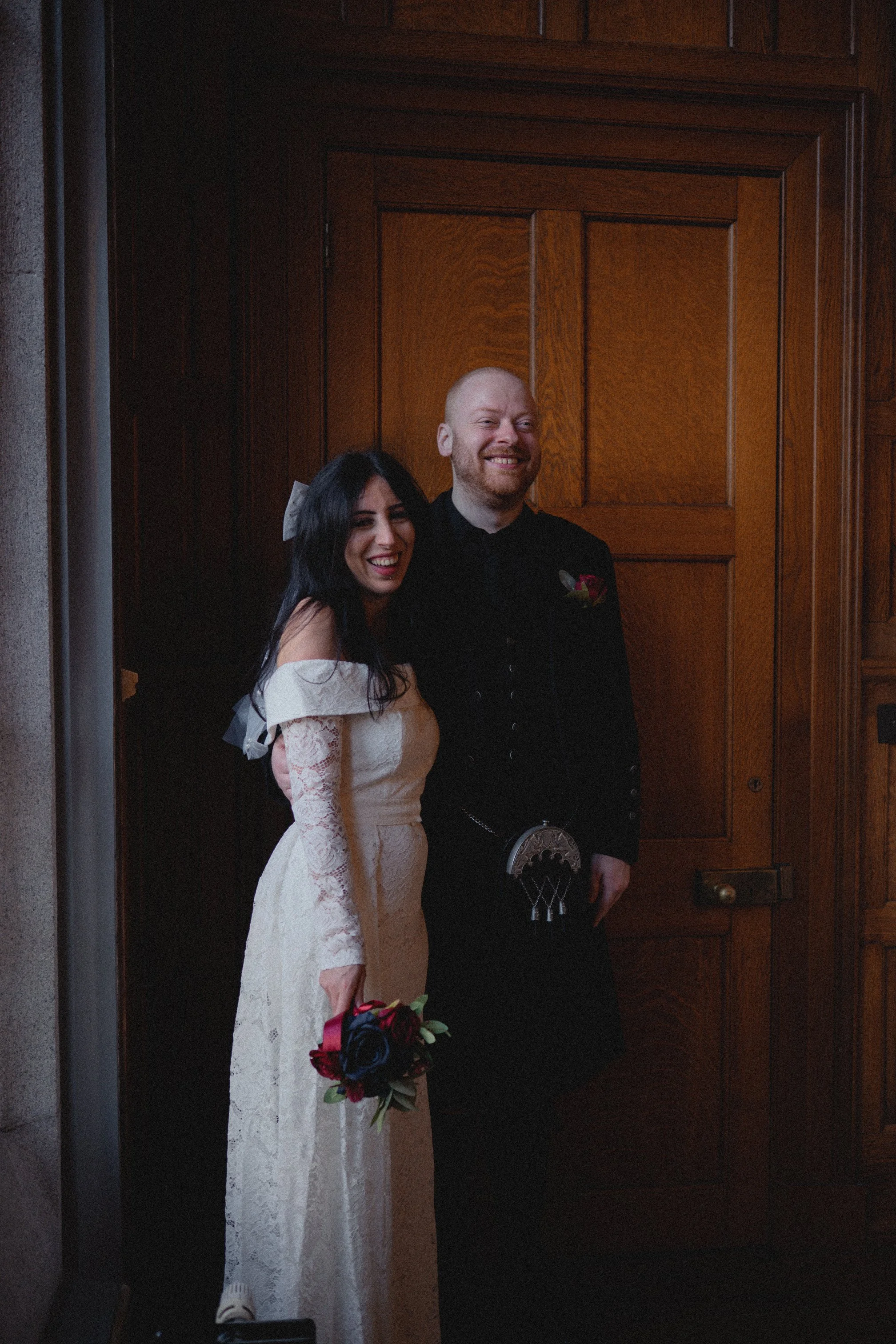 A bride and groom standing together inside a wooden-paneled room, smiling. The bride is holding a bouquet of red and black flowers and wearing an off-shoulder lace wedding dress. The groom is dressed in traditional Scottish attire with a kilt, sporra