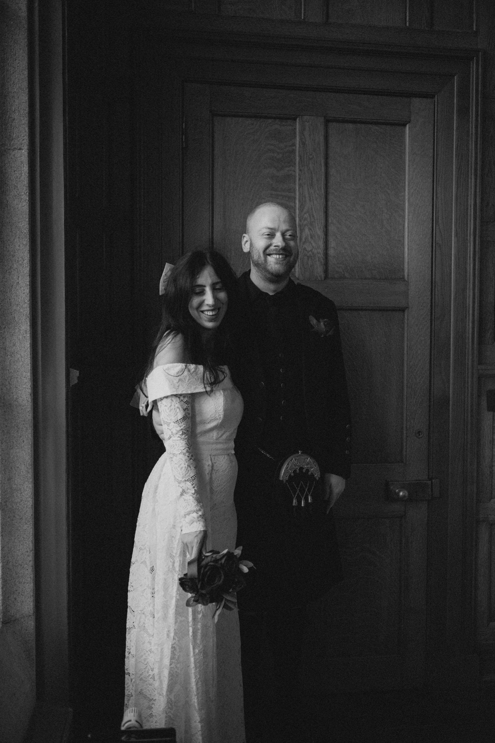 Black and white photo of a smiling bride in a lace wedding dress holding a bouquet, and a groom in a dark uniform with a decorative belt, standing in front of a wooden door.