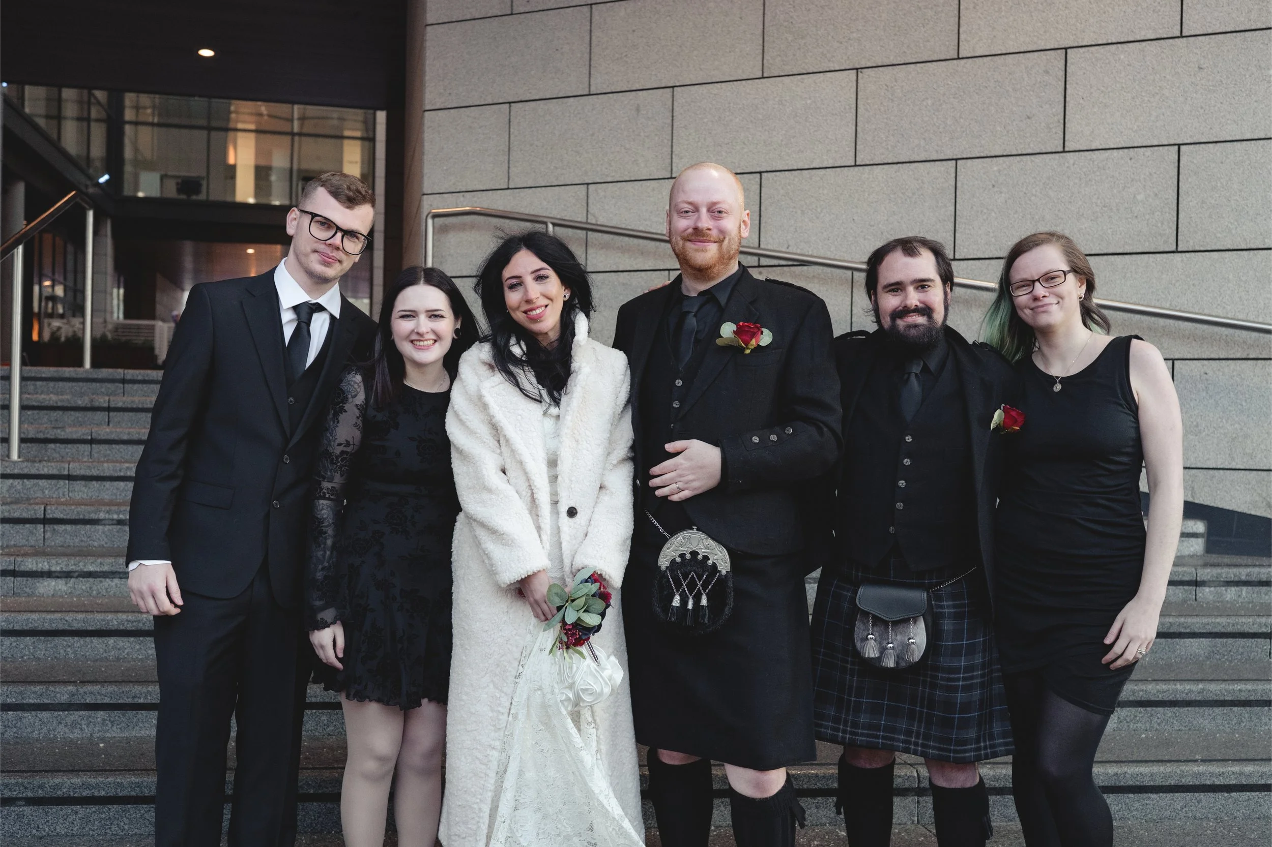 Group of six people dressed in formal attire standing on stairs outside building, smiling for photo.