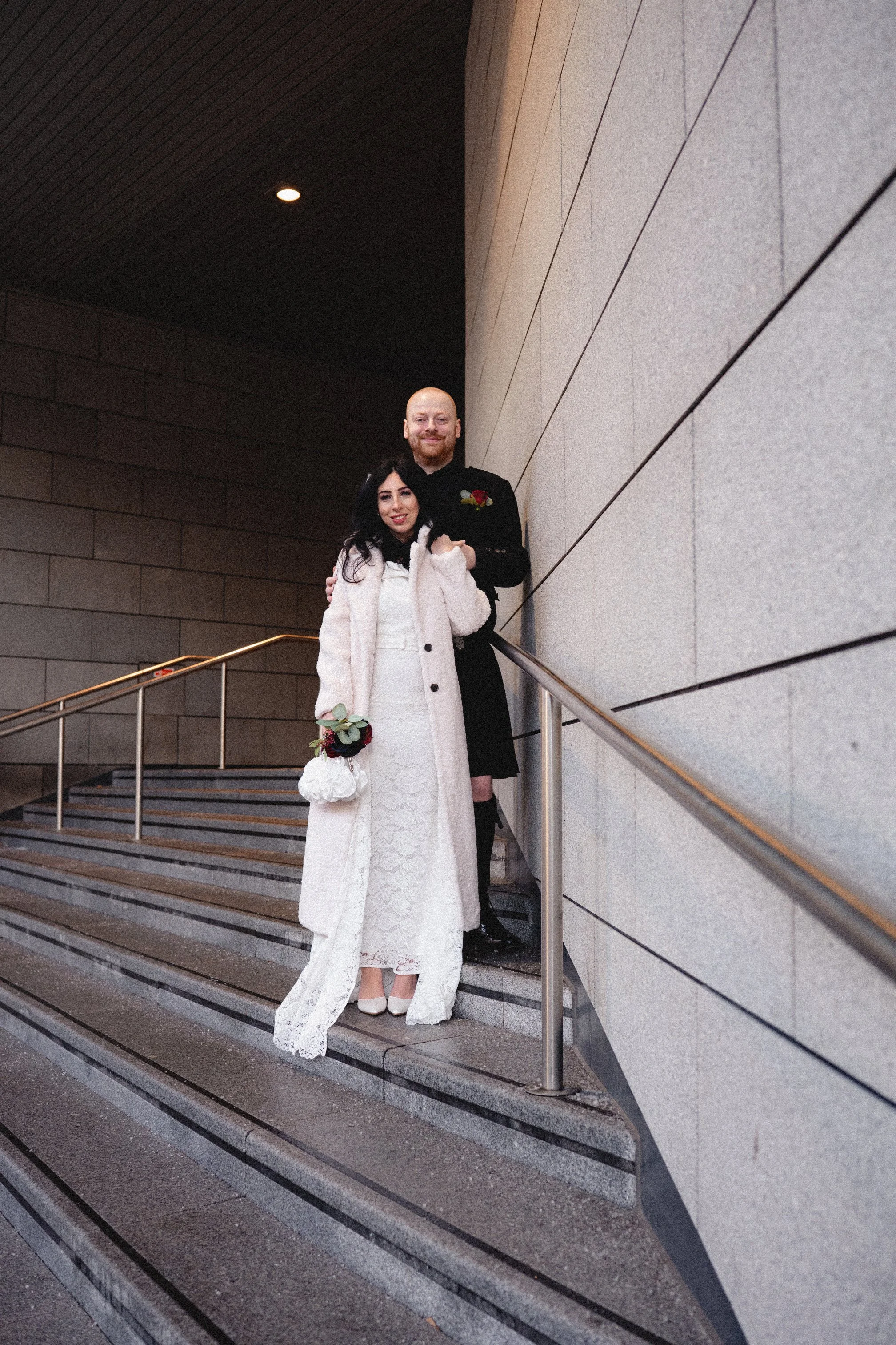 A newlywed couple standing on stairs outside a building, with the bride holding a bouquet and the groom wearing a dark suit, smiling for a photo.