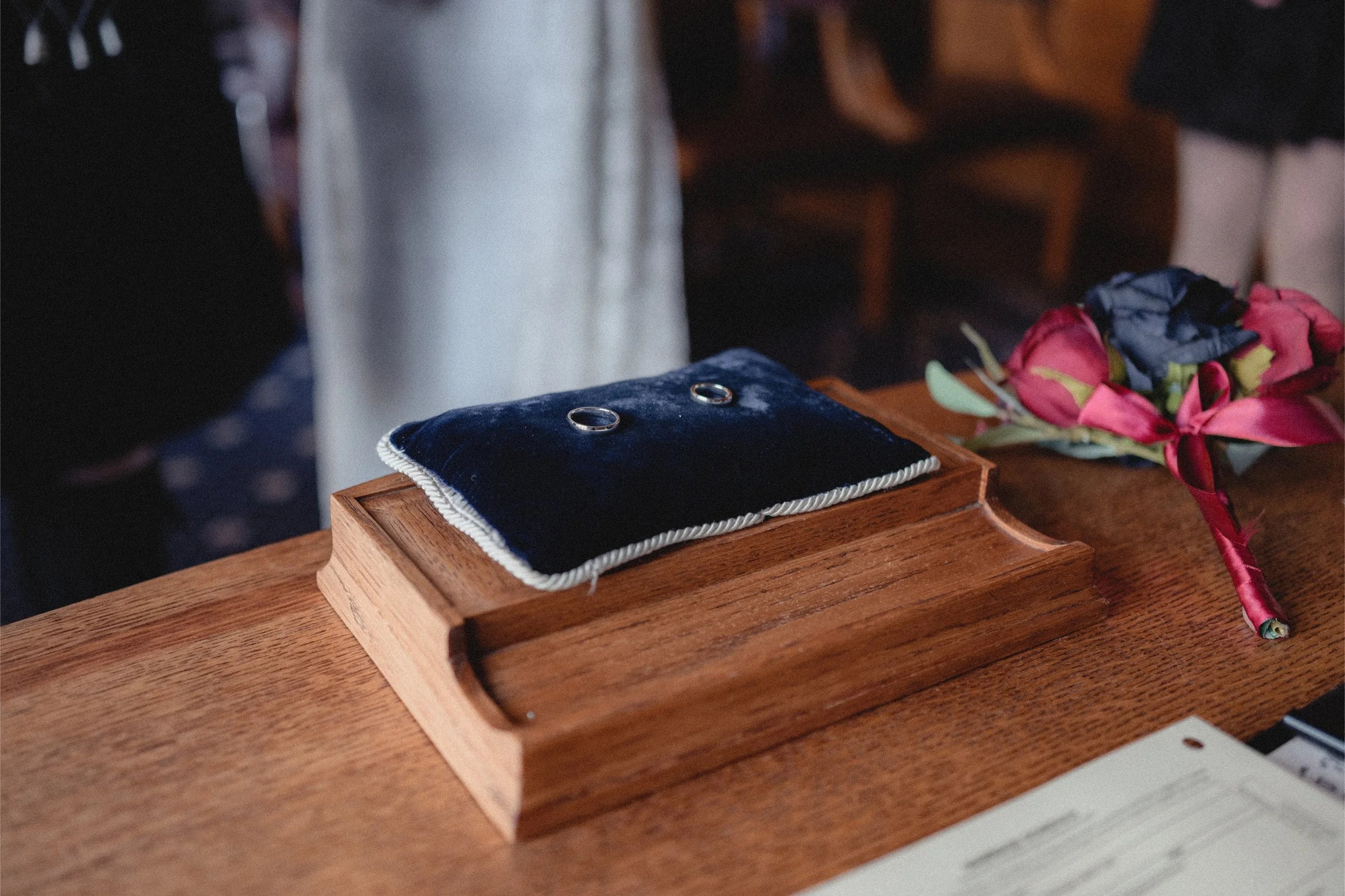 Two wedding rings on a dark blue velvet cushion on a wooden tray, with a bouquet of pink and blue flowers on a wooden table.