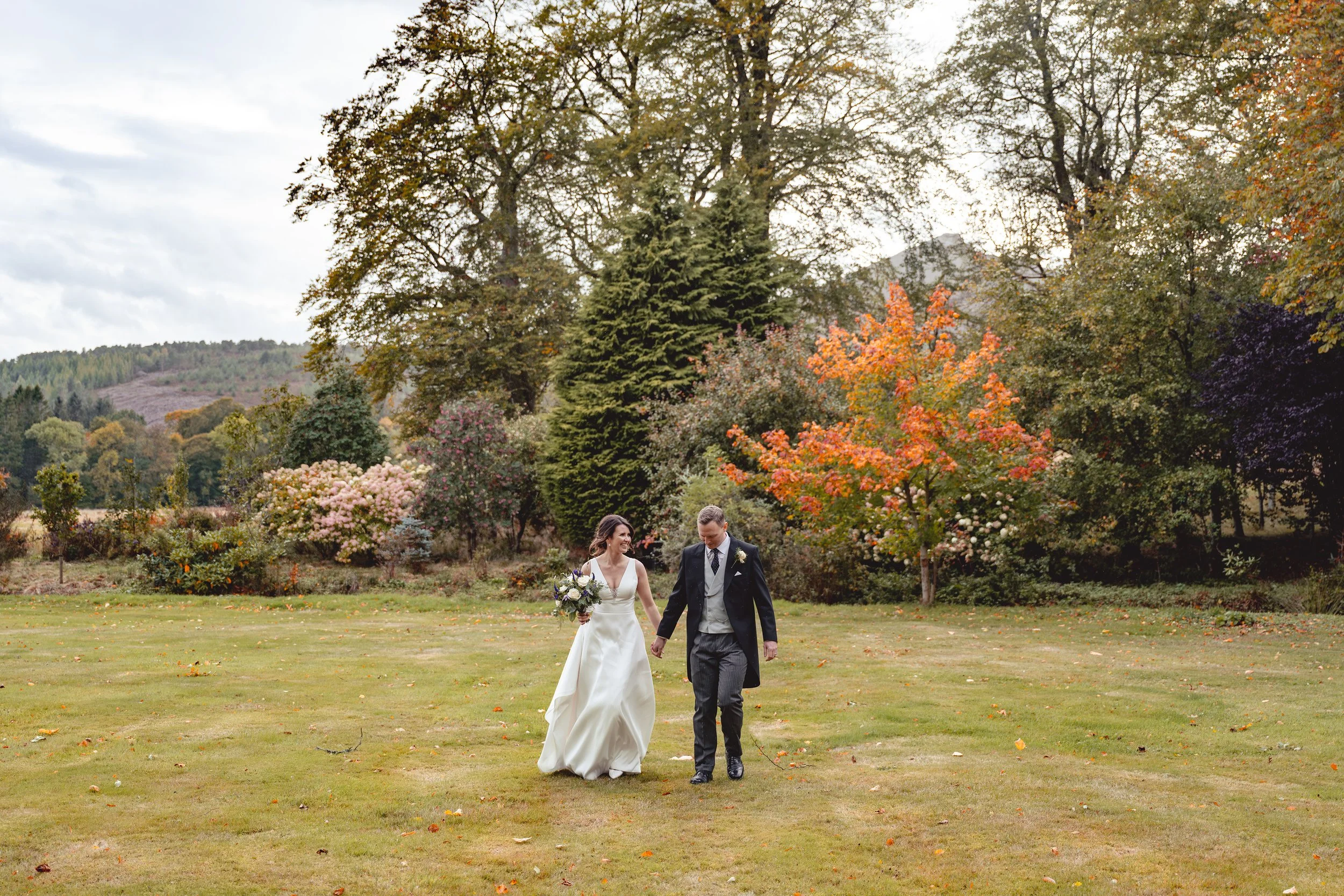 A bride and groom walking hand in hand across a grassy field during autumn, surrounded by colorful fall trees and hills in the background.
