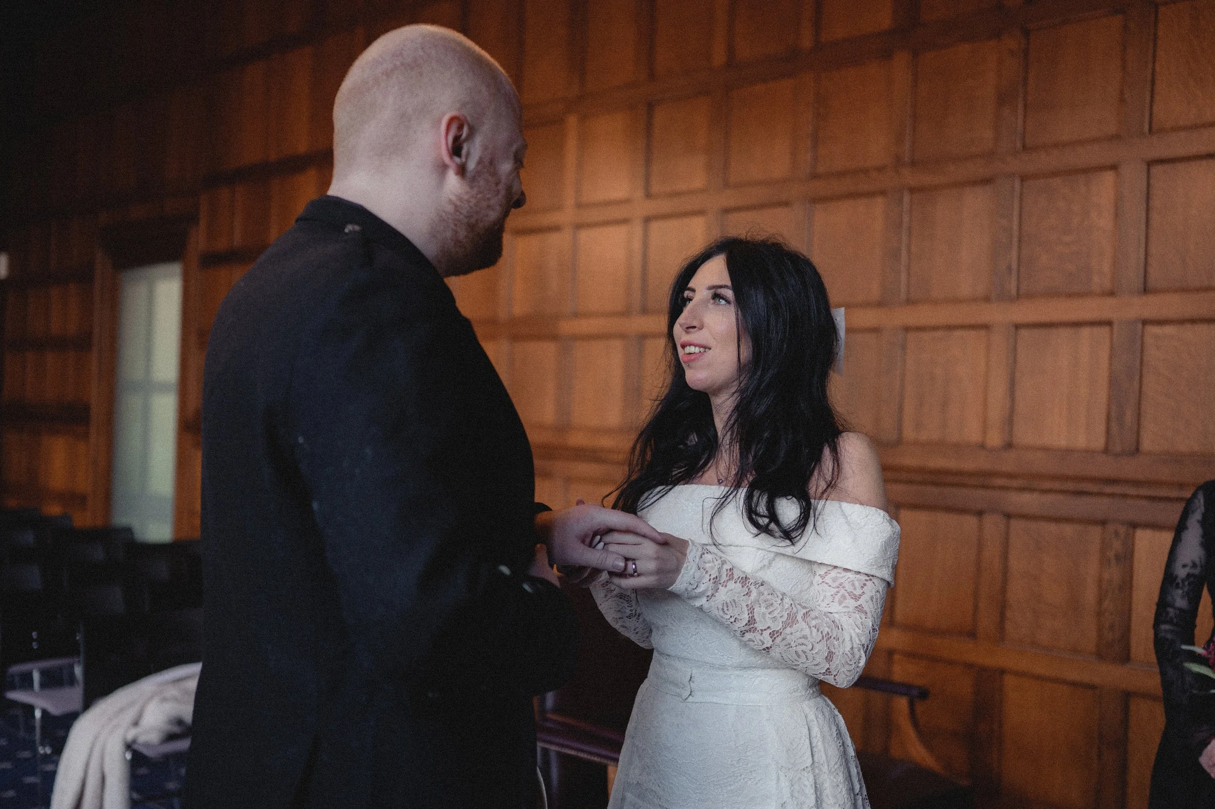 A couple holding hands, with the woman in a white lace off-shoulder dress and the man in a black suit, inside a room with wooden paneled walls.