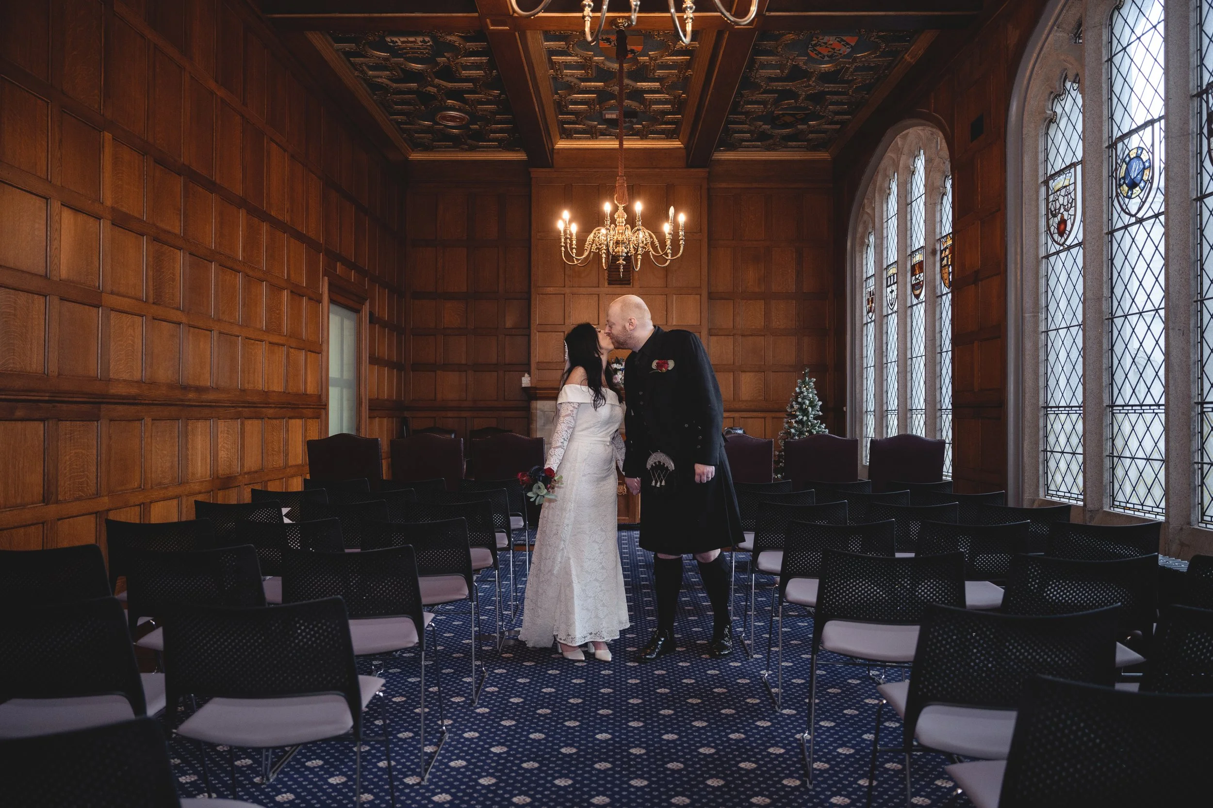 A bride and groom sharing a kiss in a decorated, wood-paneled wedding ceremony room with stained glass windows and a small Christmas tree.