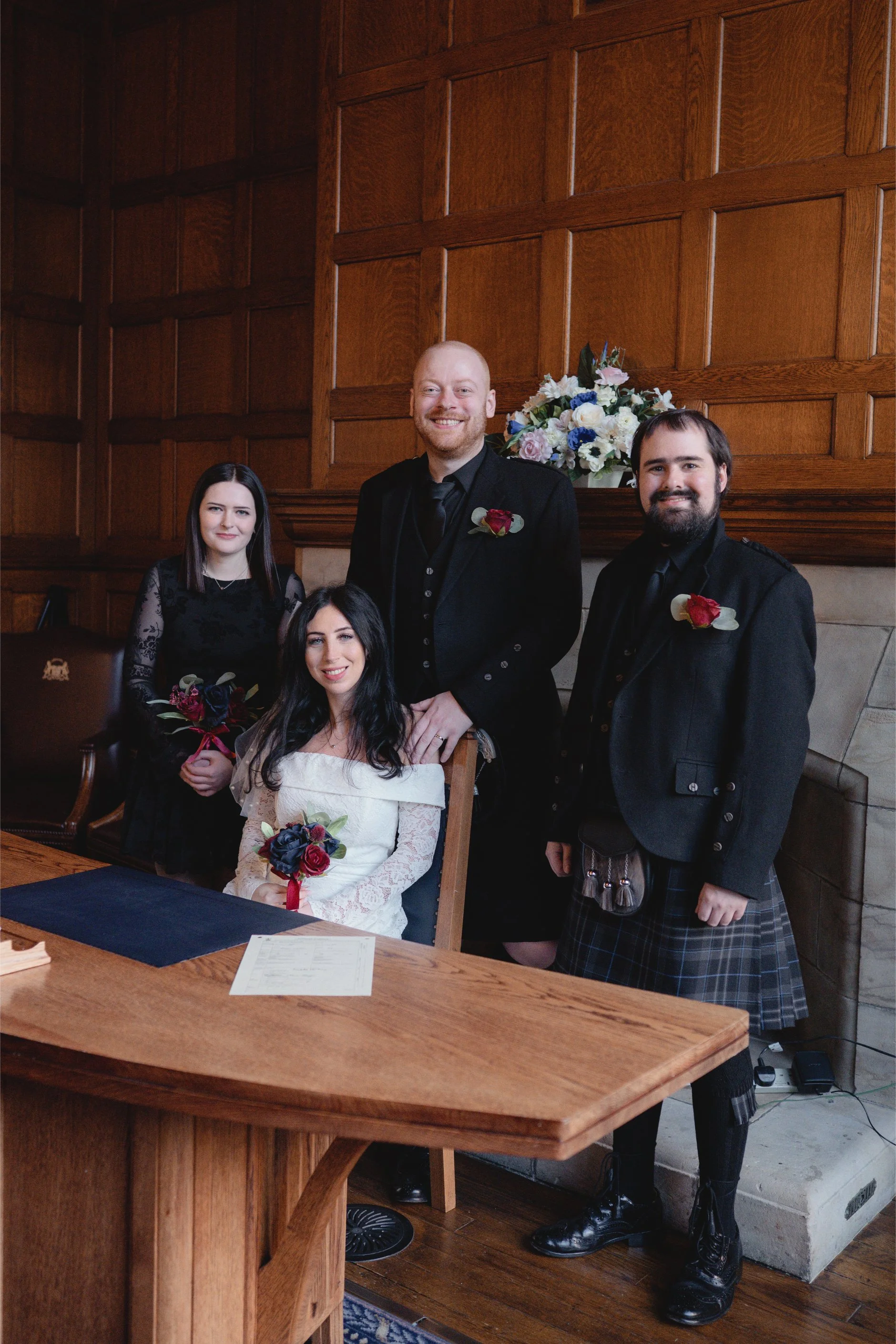 Group of five people inside a room with wooden paneling, dressed in formal attire, with three men in kilts and two women holding flower bouquets, one woman seated at a table and the others standing behind her, with floral arrangements in the backgrou