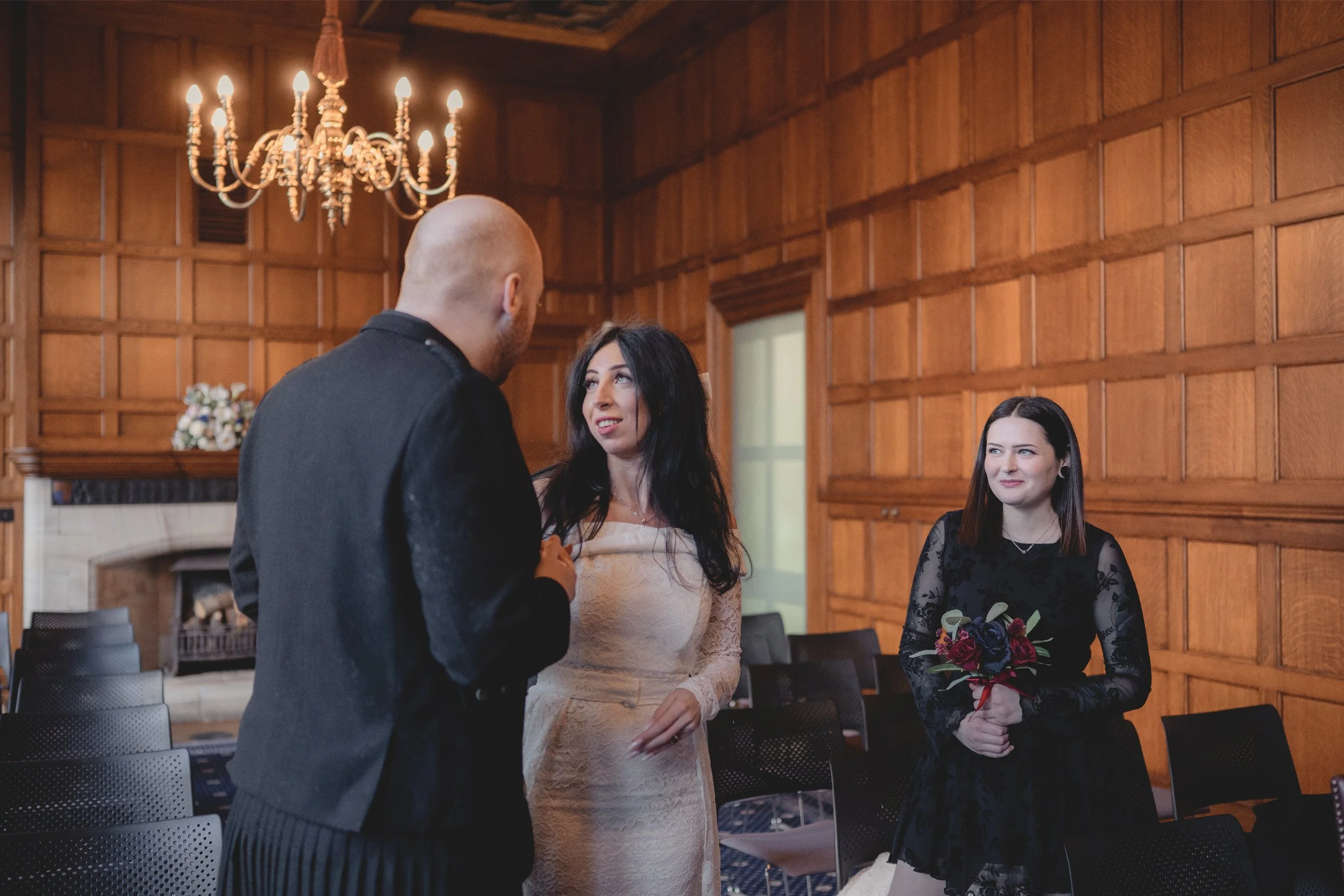A woman in a black dress holding a bouquet of roses stands in a wood-paneled room, smiling and looking at a man with a shaved head in a dark jacket, who is speaking to her. Another woman in a white lace dress stands nearby, watching the interaction.