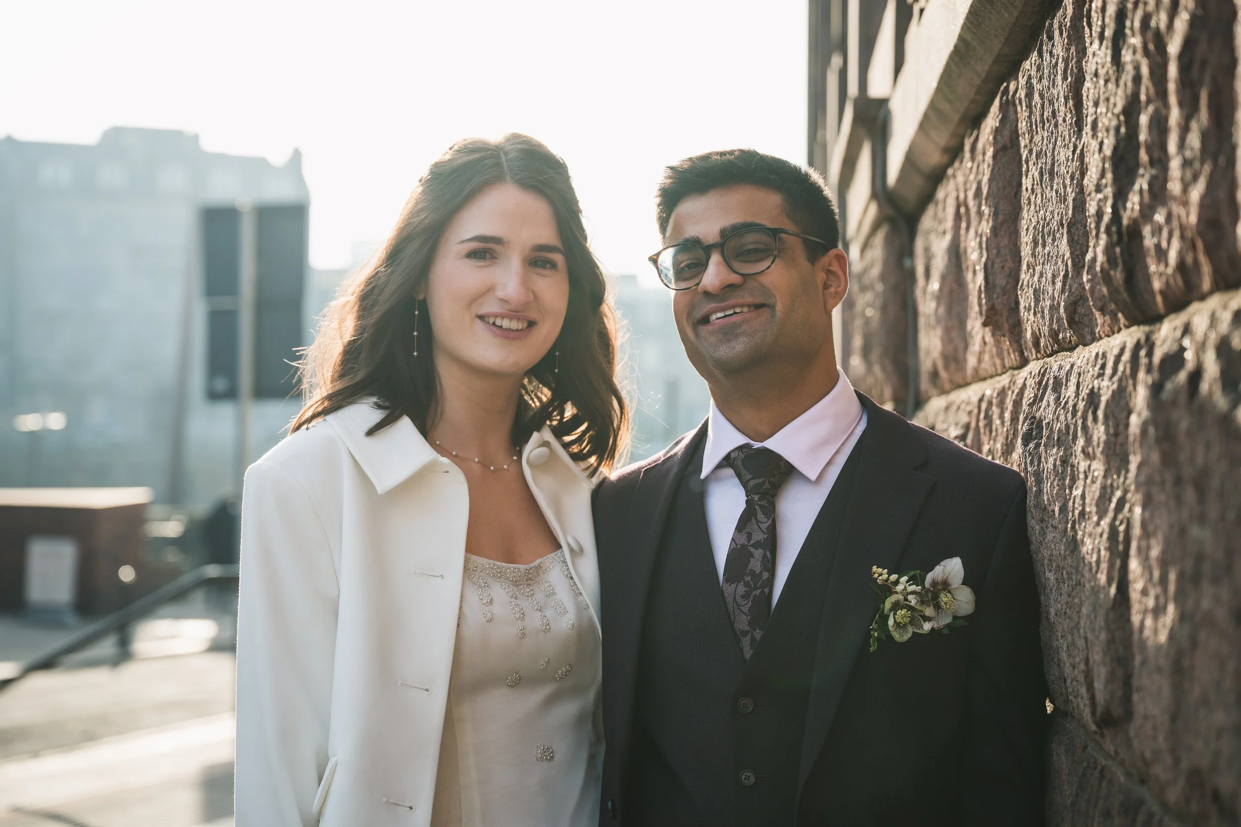 A smiling woman in a white jacket and dress standing next to a smiling man in a dark suit, tie, and glasses, leaning against a stone wall outdoors in sunlight.