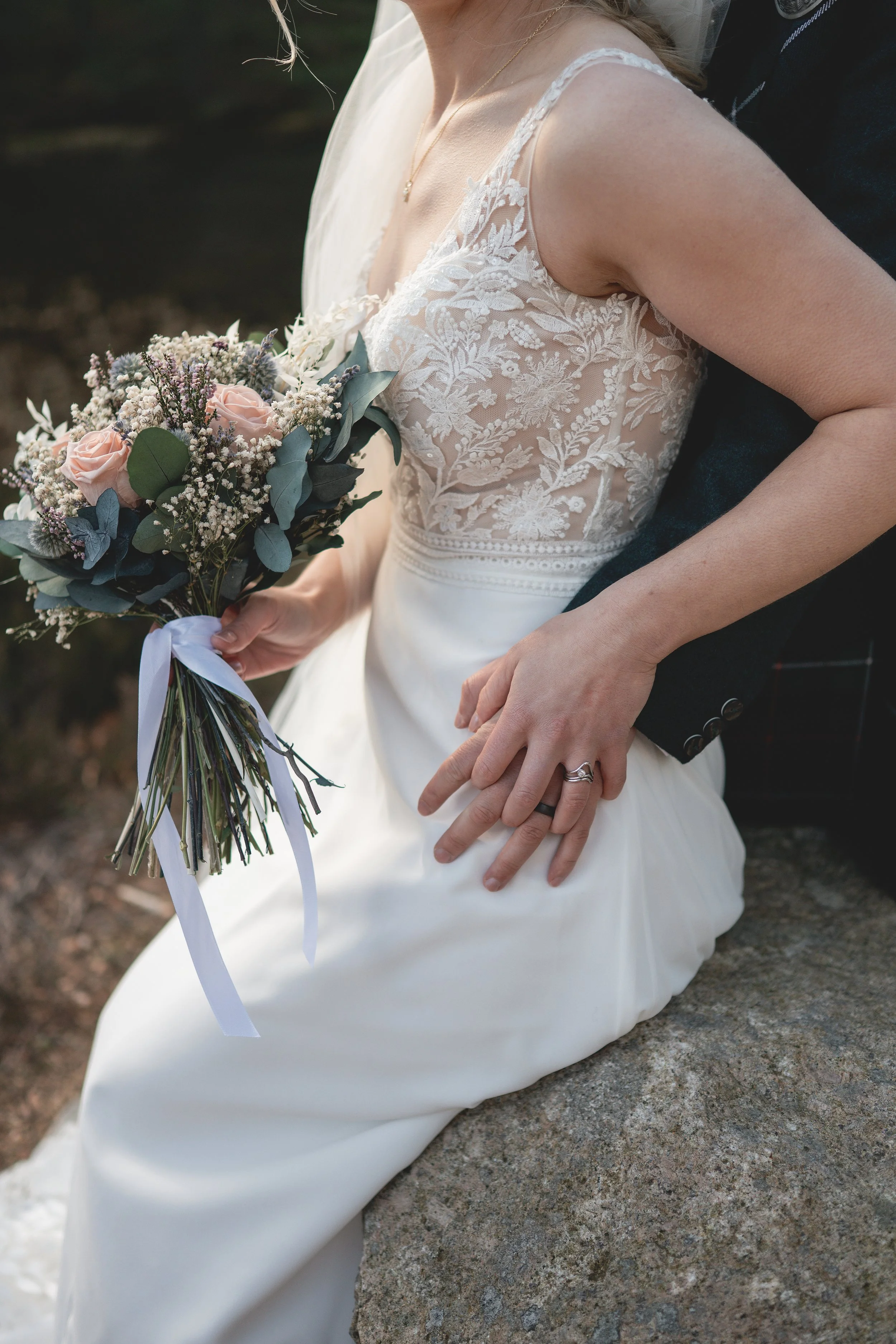 A bride in a white dress with lace detail on the top, holding a bouquet of pink roses, white baby's breath, purple thistles, and eucalyptus, sitting on a rock outdoors with a person behind her, their hand resting on her waist.