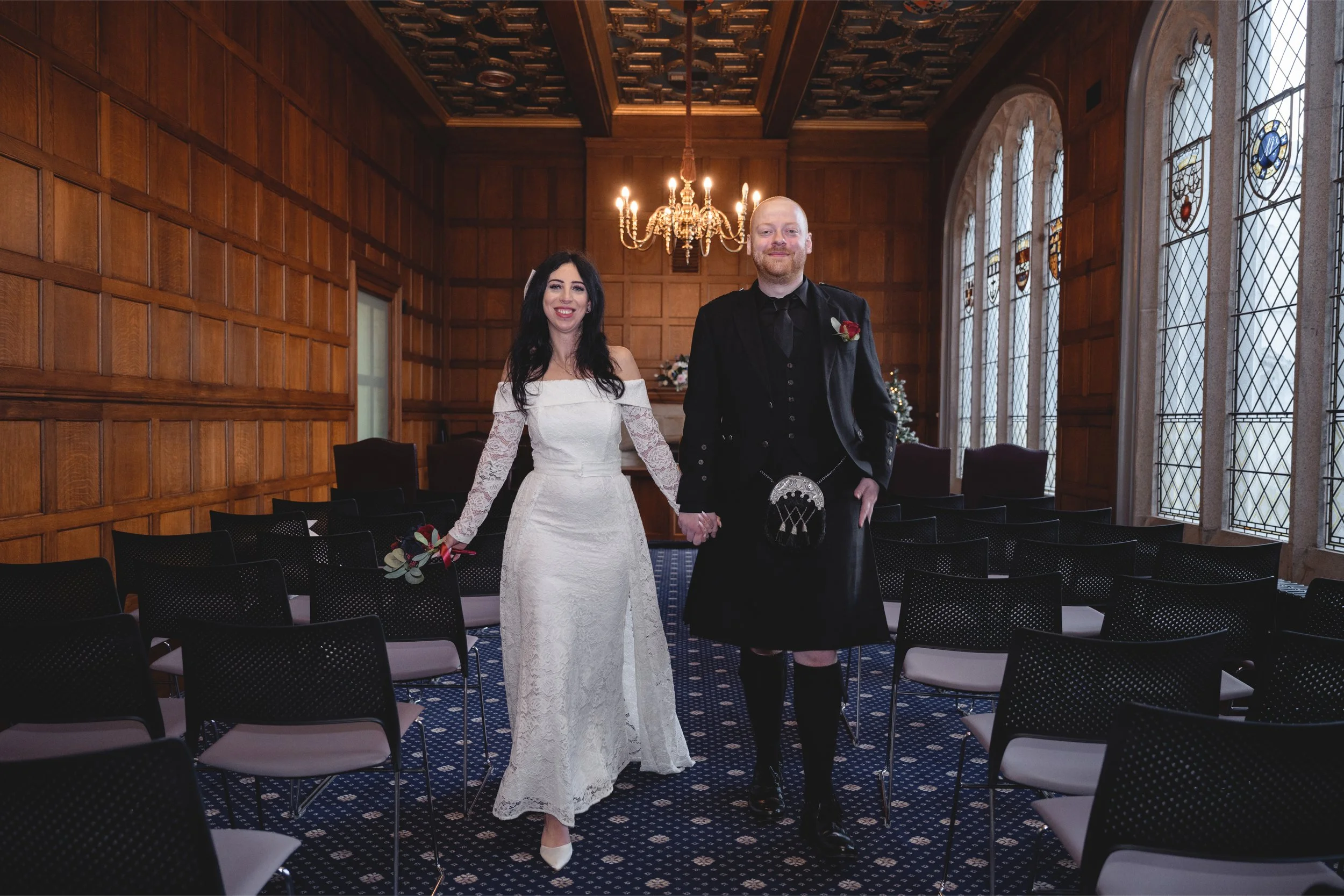A newlywed couple holding hands and smiling in a wood-paneled room with stained glass windows, a chandelier, and rows of empty chairs.