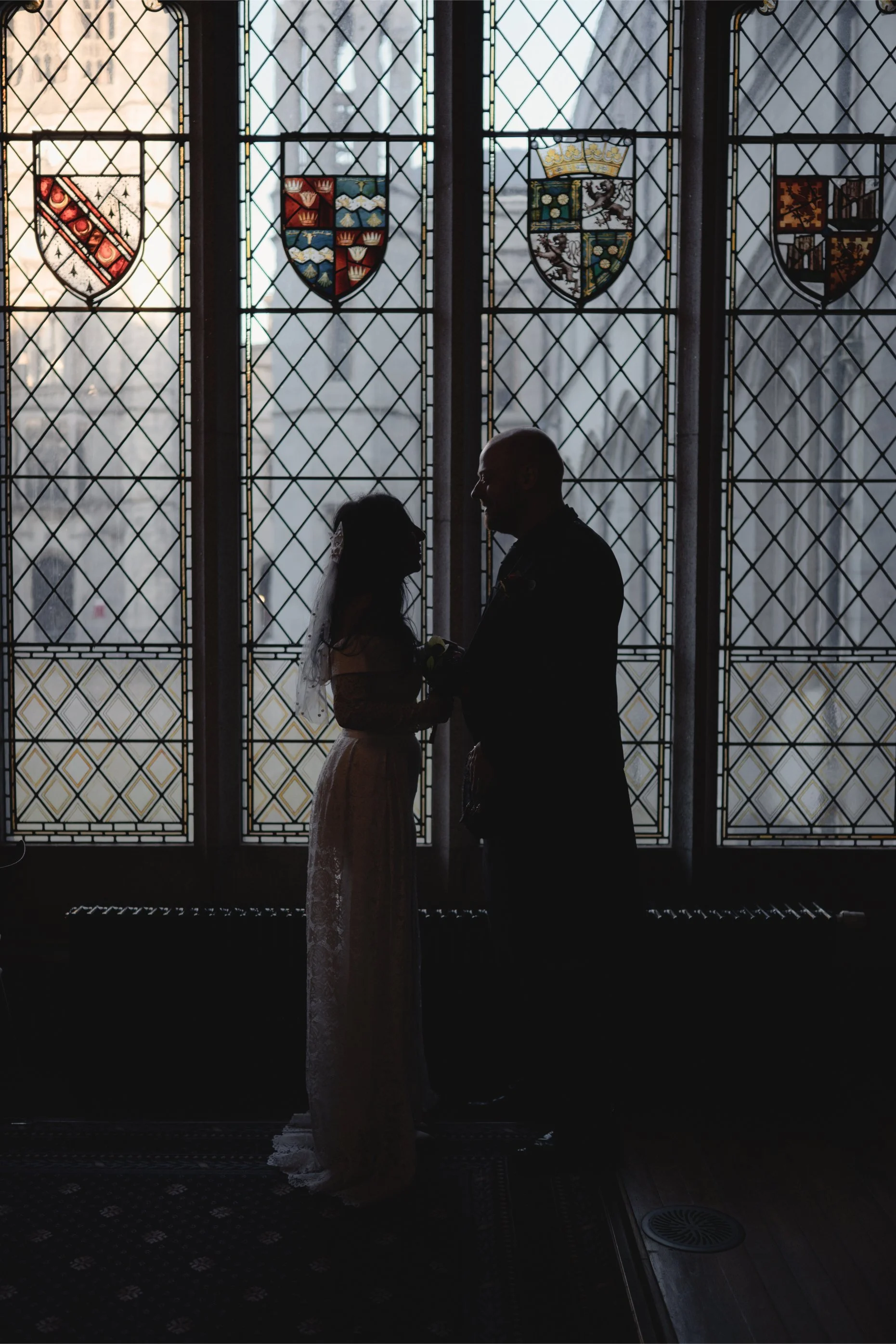 Silhouetted bride and groom holding hands in front of stained glass windows with heraldic shields.
