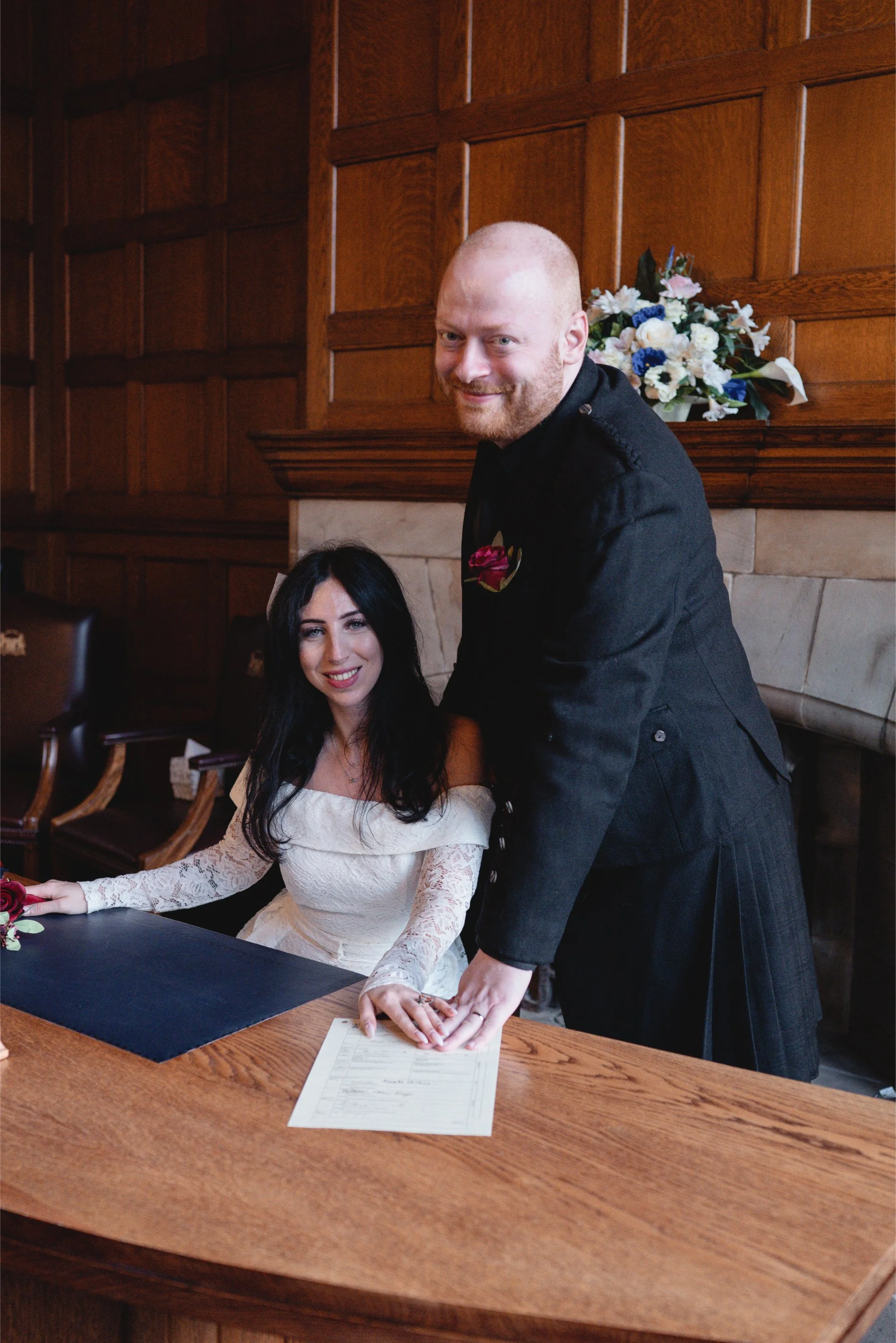 A newlywed couple signing their marriage certificate at a wooden table in a room with dark wood paneling and a bouquet of flowers in the background.
