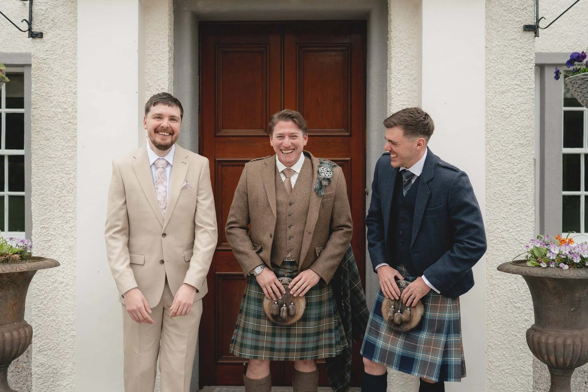 Three men standing in front of a wooden door, two wearing kilts and suits, and one in a beige suit, all smiling and laughing.