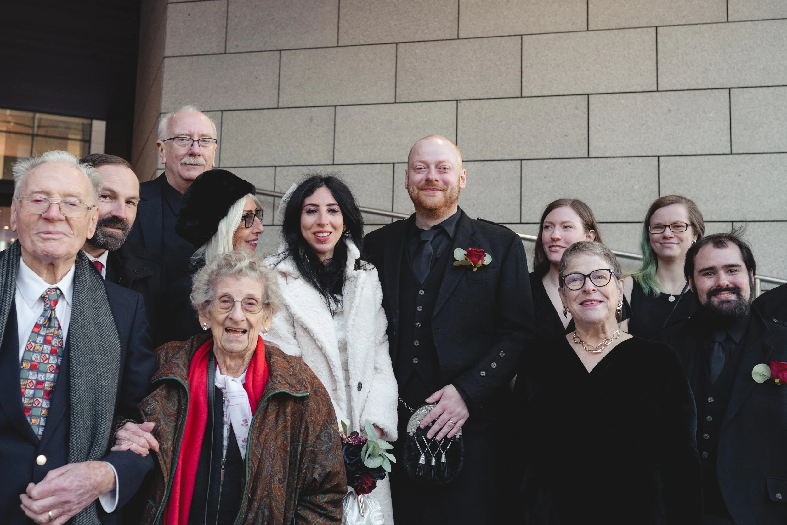 A group of people dressed in formal attire, including wedding clothing, standing together outdoors against a brick wall. The group includes elderly individuals, middle-aged adults, and young adults smiling at the camera.