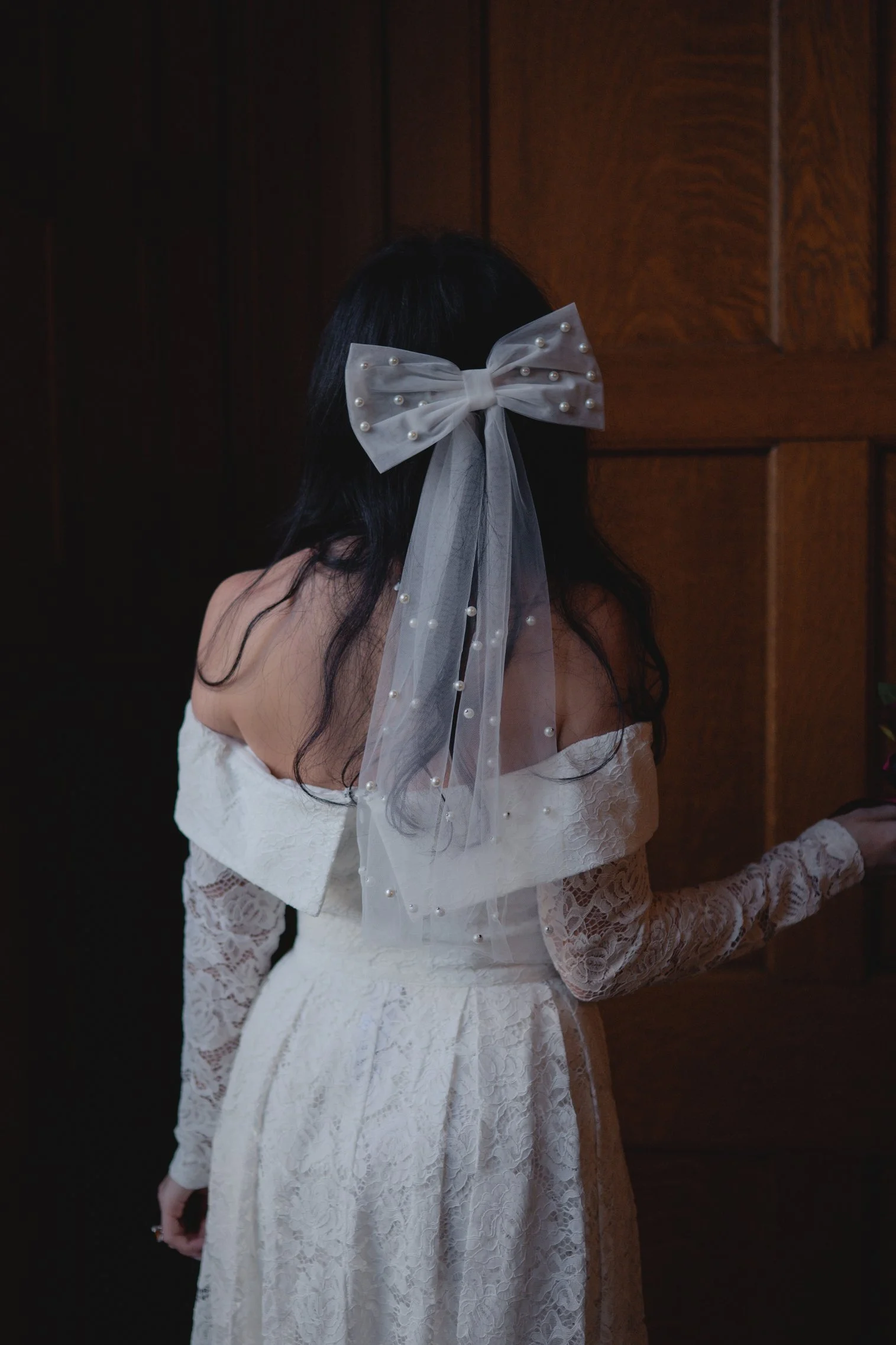 Woman in a white lace dress with off-shoulder neckline, facing a wooden wall, wearing a pearl-embellished sheer bow and veil in her dark hair.