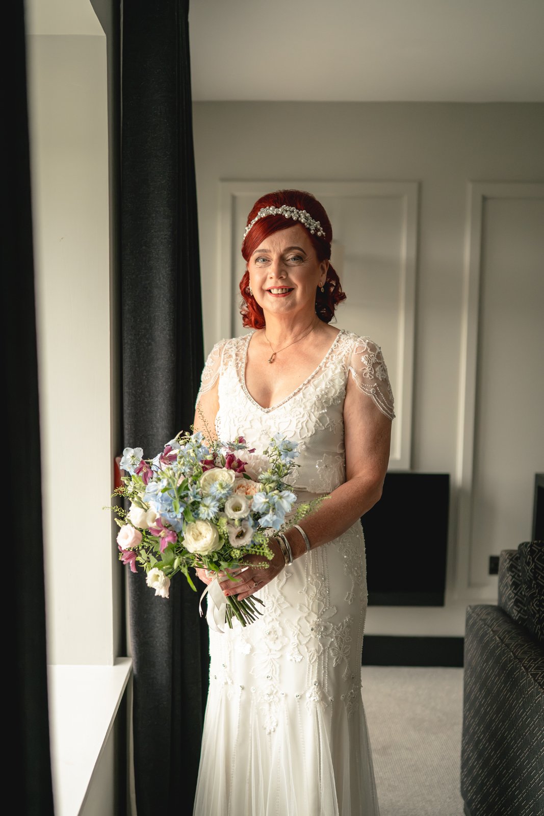 A smiling woman in a white wedding dress holding a bouquet of flowers stands indoors near a window.