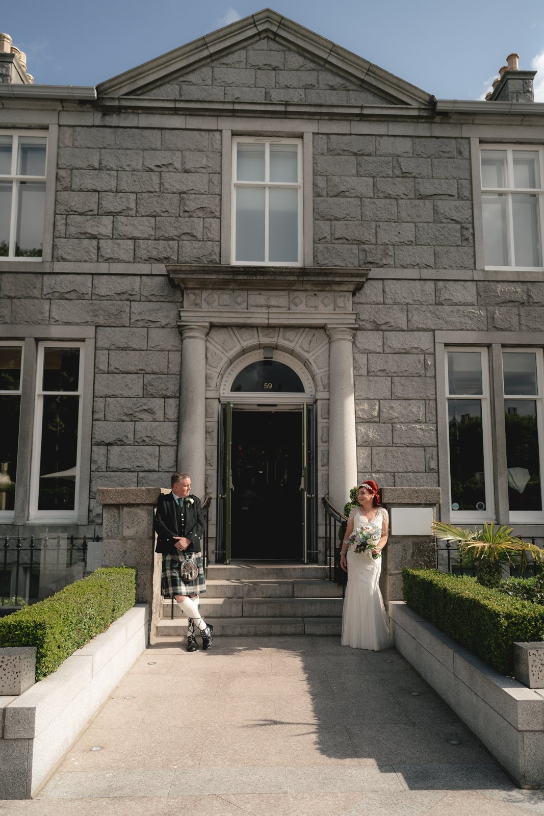 Bride and groom standing outside a large stone building, possibly a wedding venue, with the groom dressed in traditional Scottish kilt attire and the bride in a white wedding gown holding a bouquet.