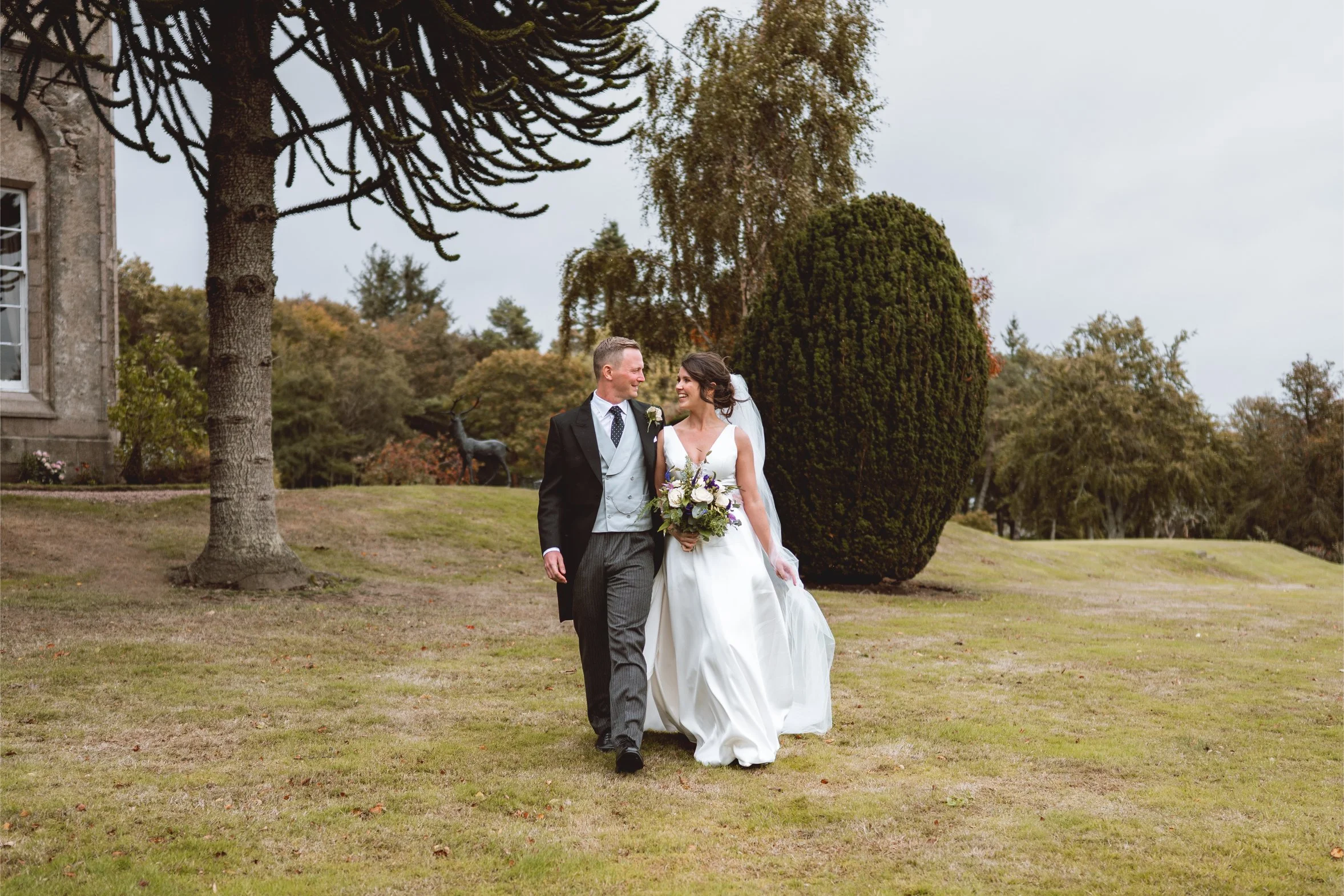 A bride and groom walk arm in arm outdoors, smiling at each other on their wedding day. The bride wears a white gown and veil, holds a bouquet, and the groom wears a black coat, gray vest, and striped trousers. The background features trees, neatly trimmed bushes, and an overcast sky.