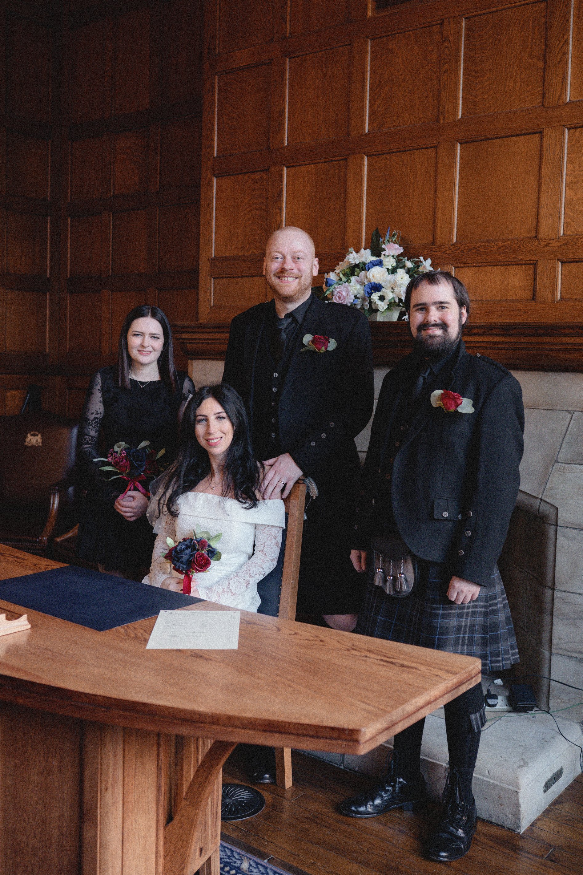 Group of four people in formal attire at a wedding, standing and sitting in a wood-paneled room with a flower arrangement behind them.
