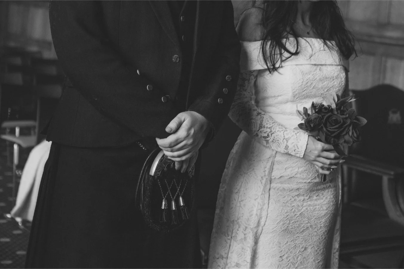 Black and white photo of a couple at a wedding or formal event. The woman is holding a bouquet of roses, and the man is dressed in formal attire. Only the torsos and hands are visible, with the man's hands clasped in front of him and the woman holdin
