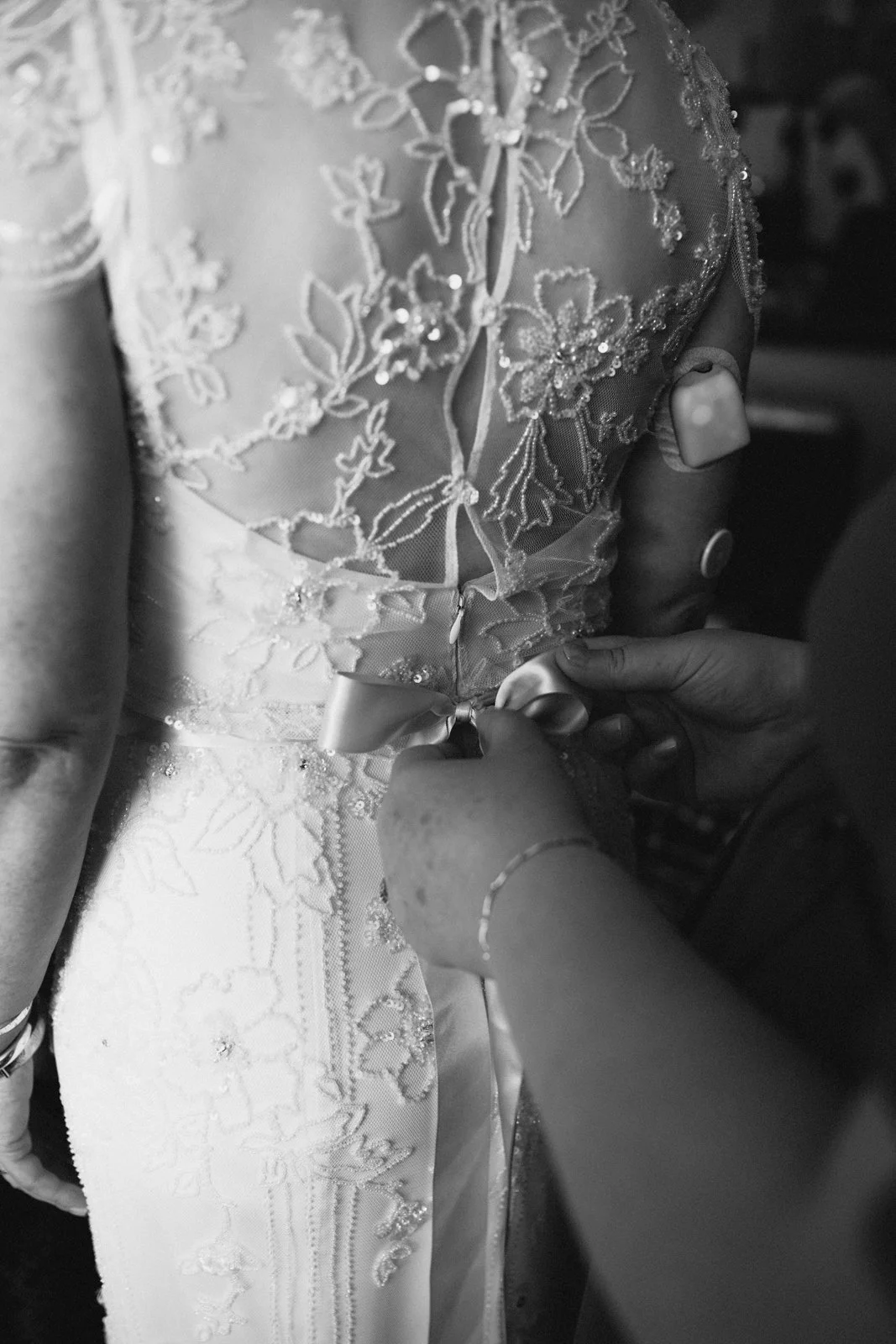 Close-up of a wedding dress with intricate lace embroidery, as someone fastens a satin bow at the back.
