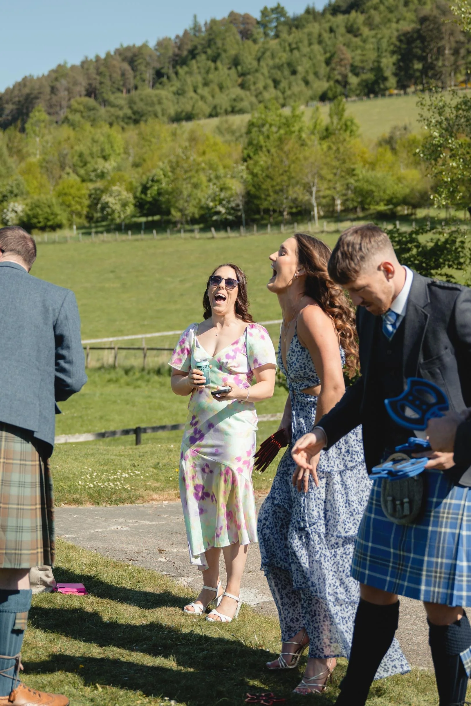 A group of people outdoors, laughing and enjoying themselves. Two women are wearing summery dresses, and two men are dressed in traditional Scottish kilts. The background features a lush green hillside and a bright, sunny sky.