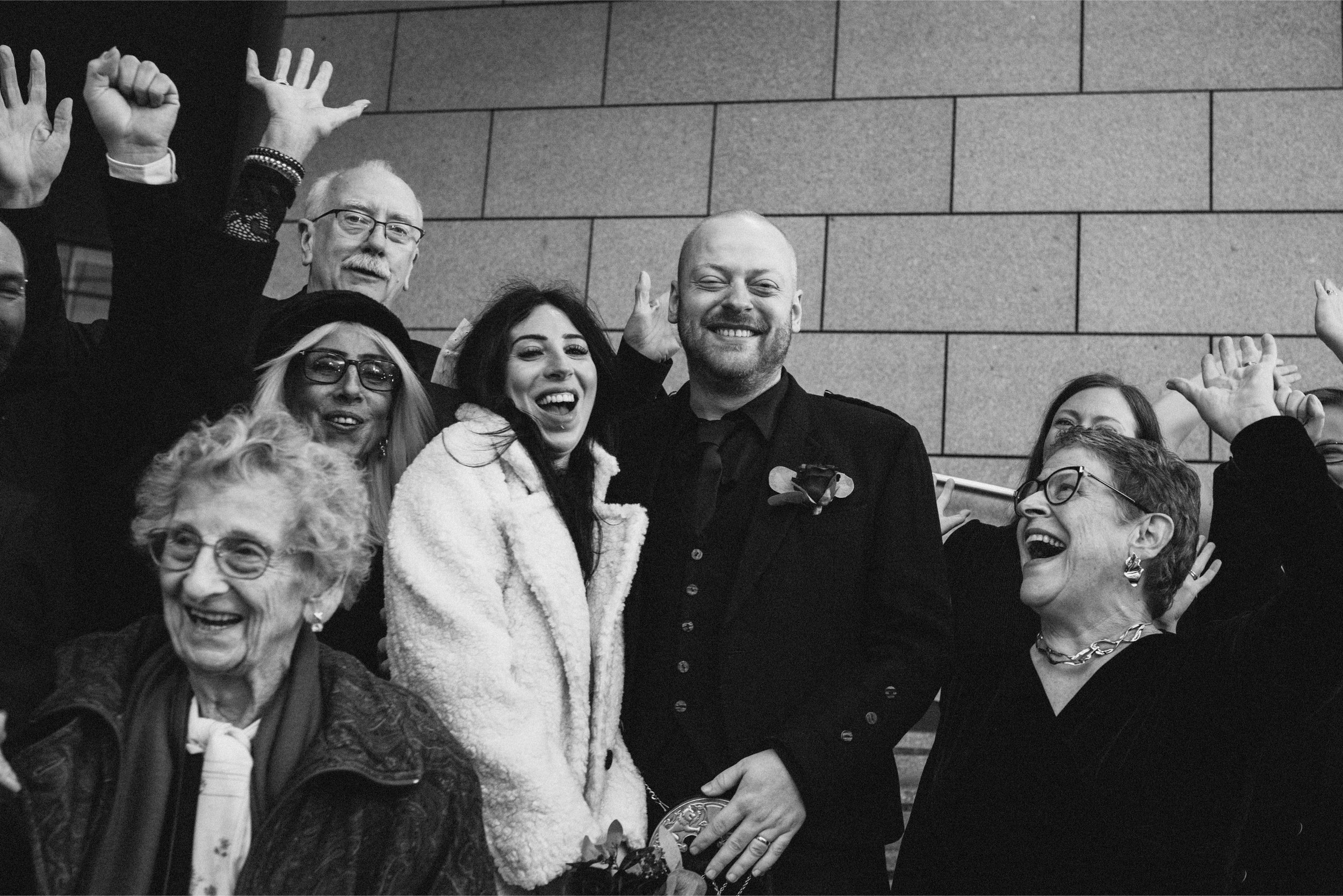 Group of diverse people celebrating and laughing together indoors, with some raising their hands, in front of a tiled wall.