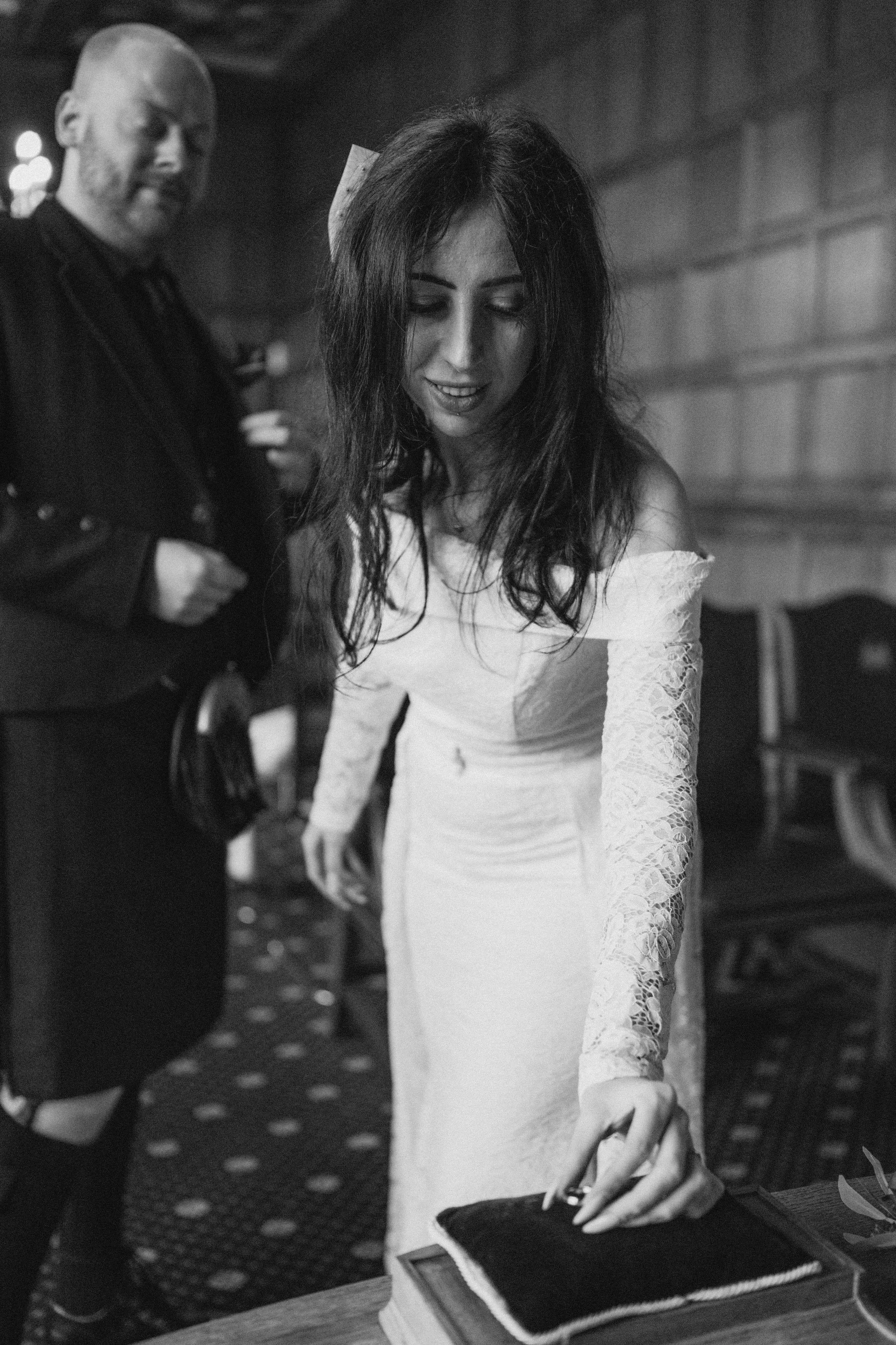 A woman in a white lace dress with long sleeves standing at a table, touching a book or box with her right hand, while a man with a beard in a dark outfit stands behind her, in a dimly lit room with wood-paneled walls.