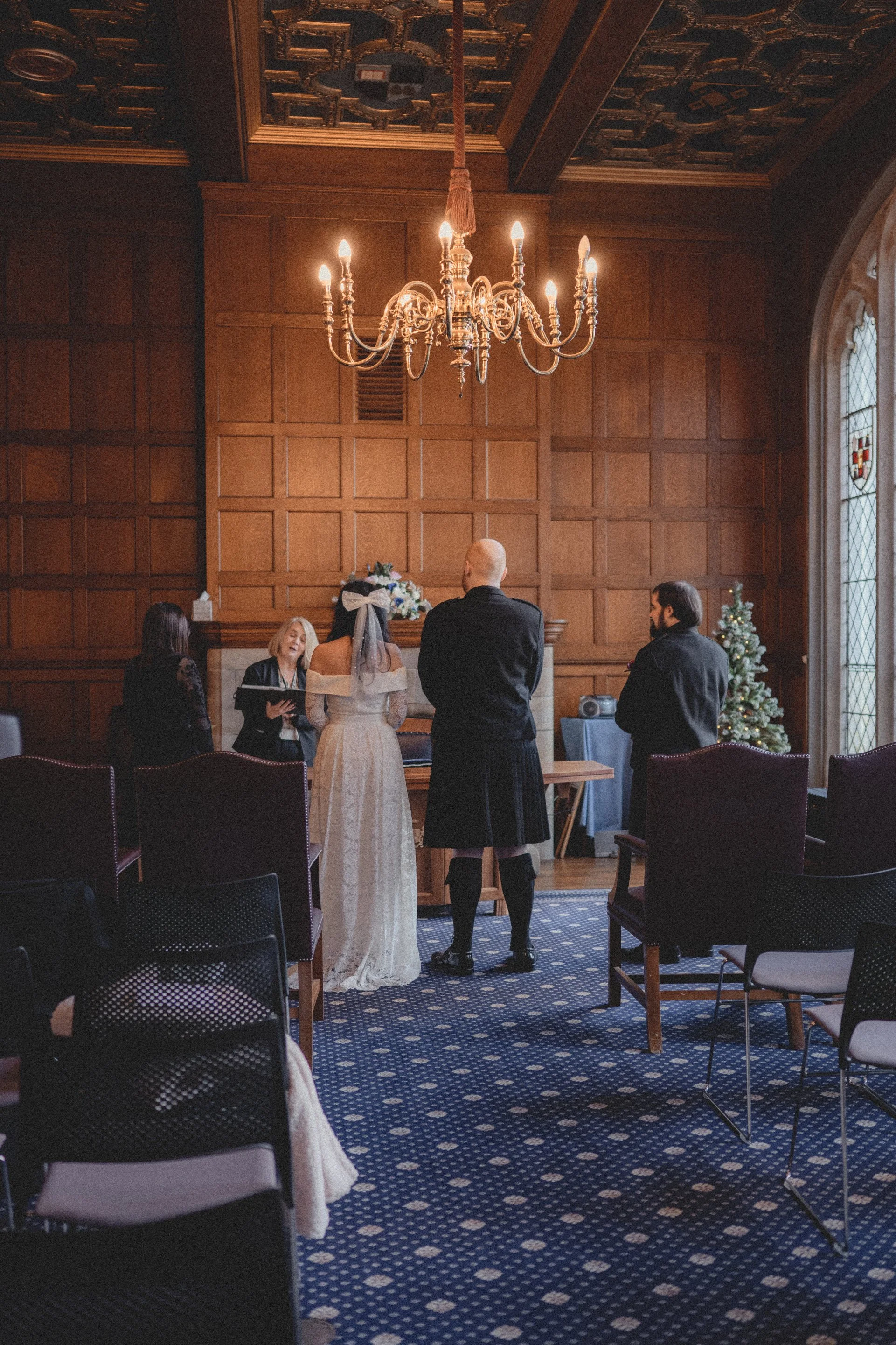 A couple getting married in a wood-paneled room with a chandelier, Christmas tree, and stained glass window, with officiant and witness present.