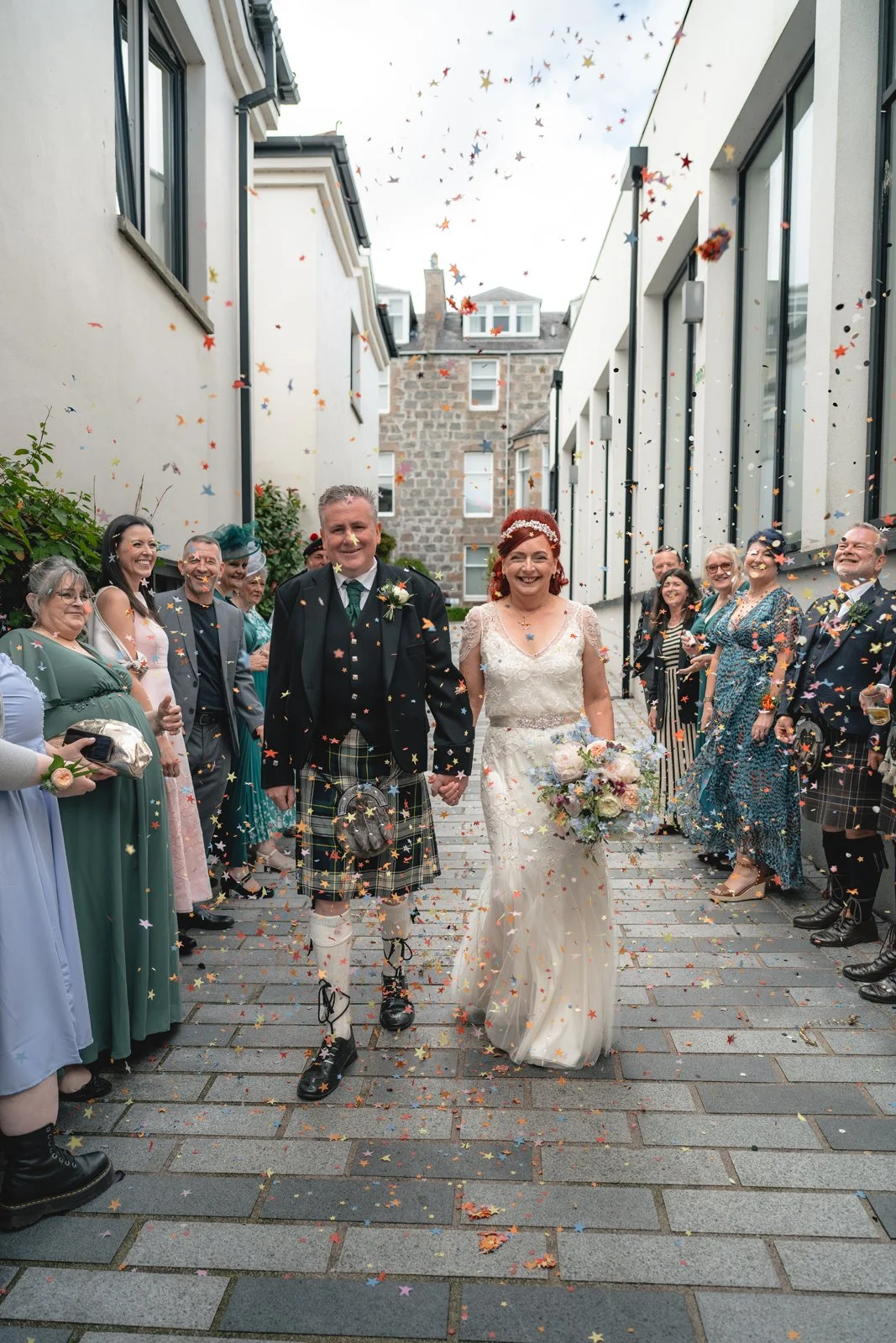 A newly married couple walking hand-in-hand through a shower of colorful confetti, surrounded by friends and family in an outdoor urban setting.