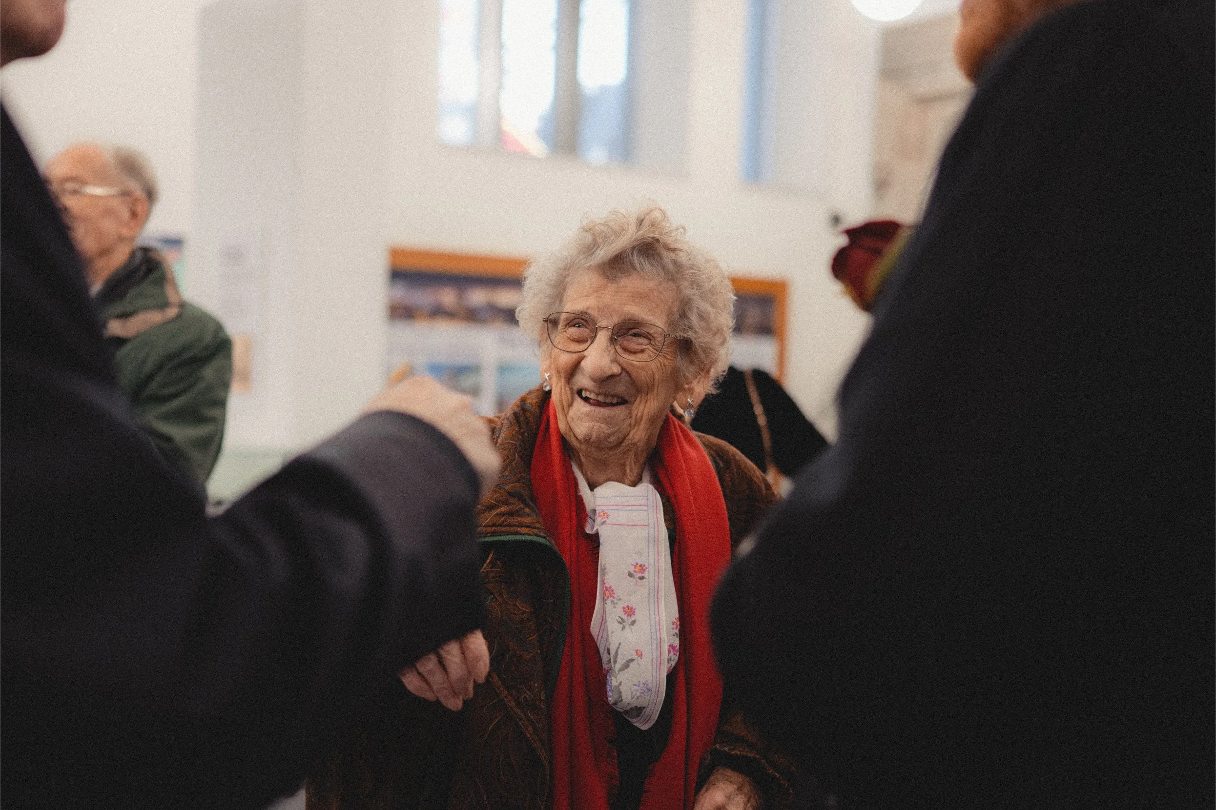 An elderly woman with gray curly hair and glasses smiling, surrounded by people in a social setting, indoors with bright natural light.