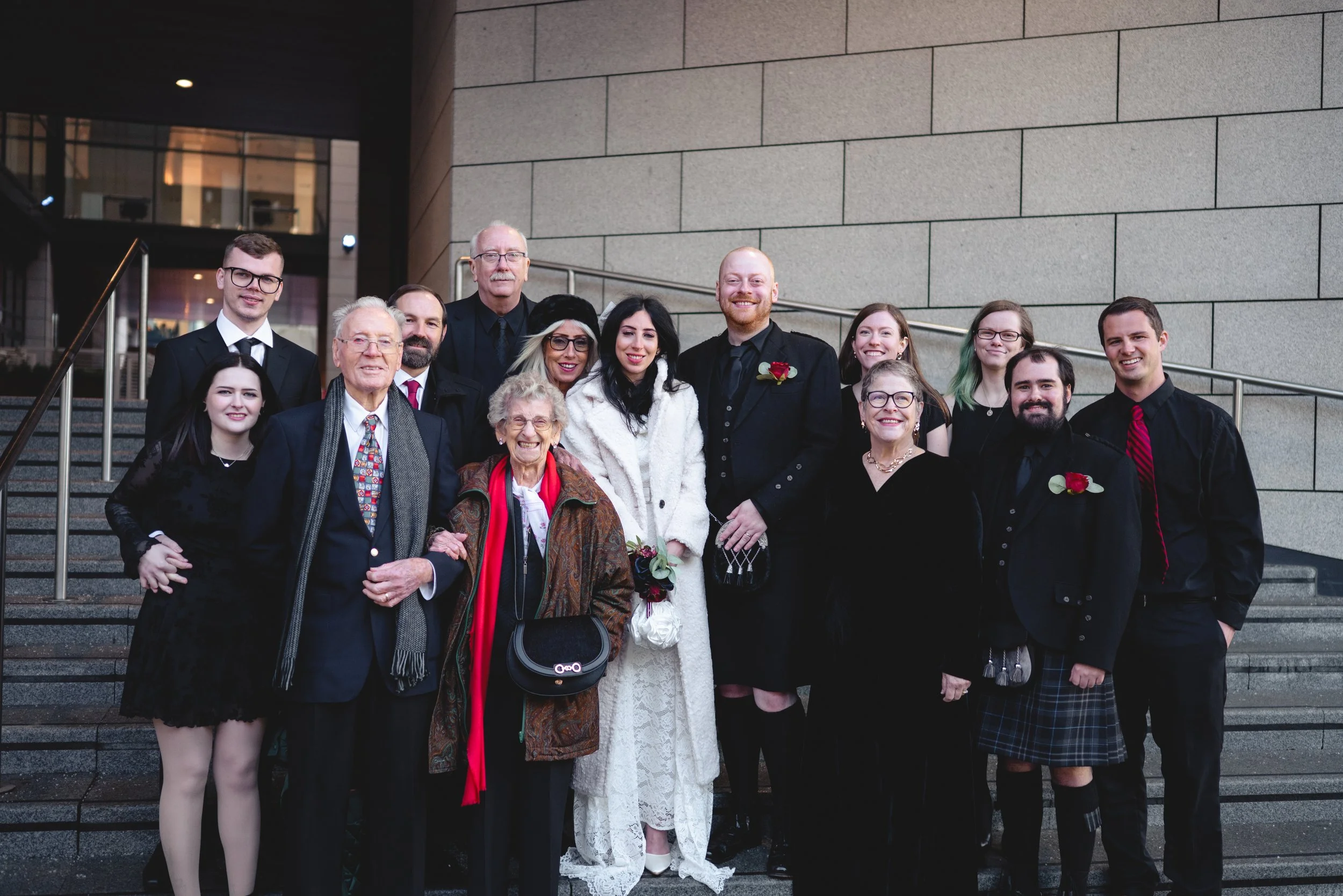 Group of people dressed in formal attire standing on outdoor steps for a wedding photo.
