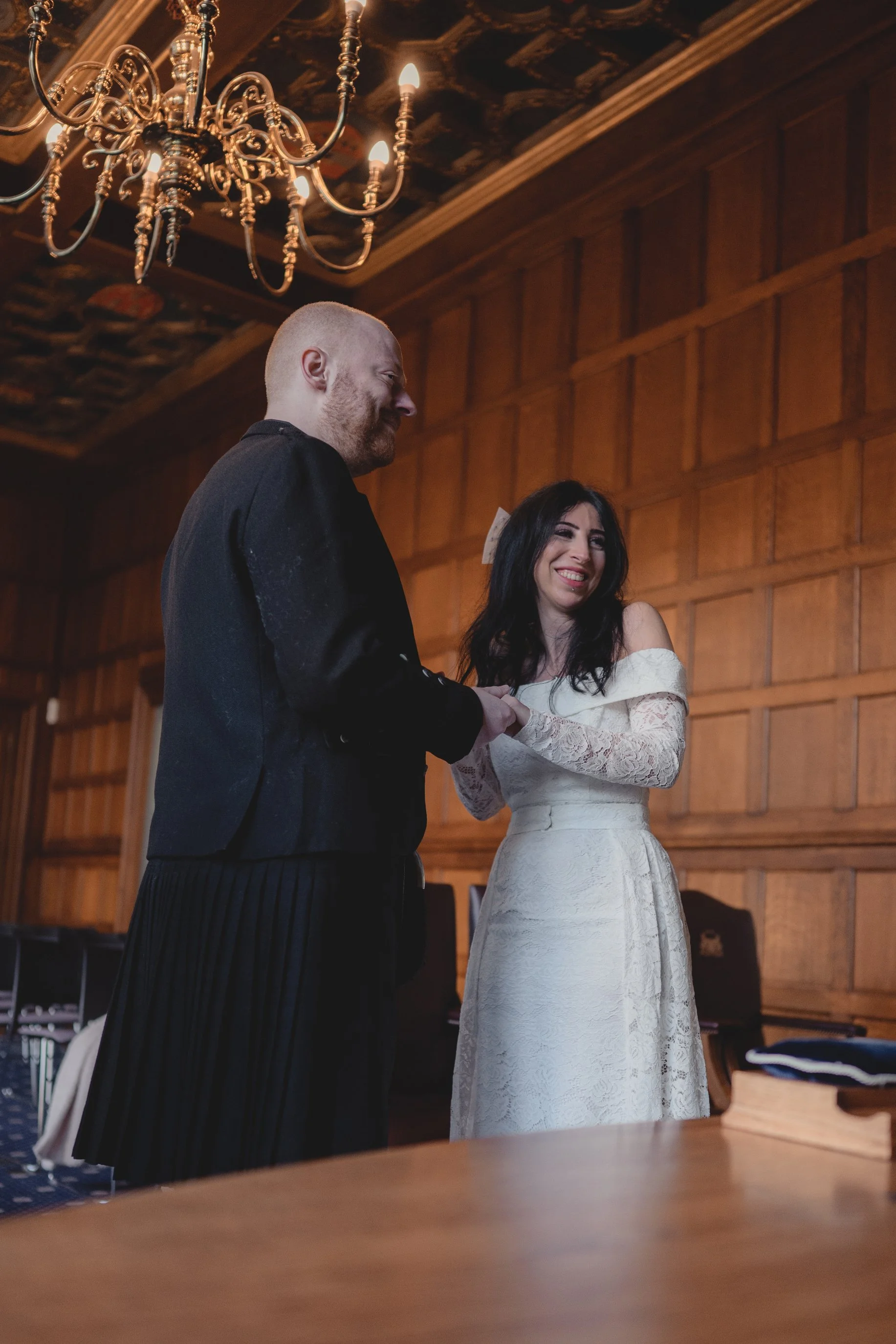 A man and woman holding hands and smiling during a wedding ceremony in a wood-paneled room with a chandelier.