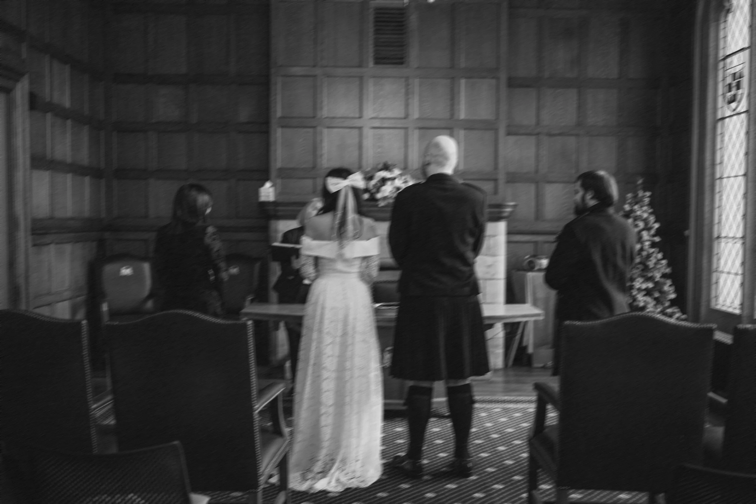 A wedding ceremony with a bride in a white dress and veil, a man in a kilt, and two women, one with a face mask, standing in front of a wooden-paneled wall with a fireplace and flowers.