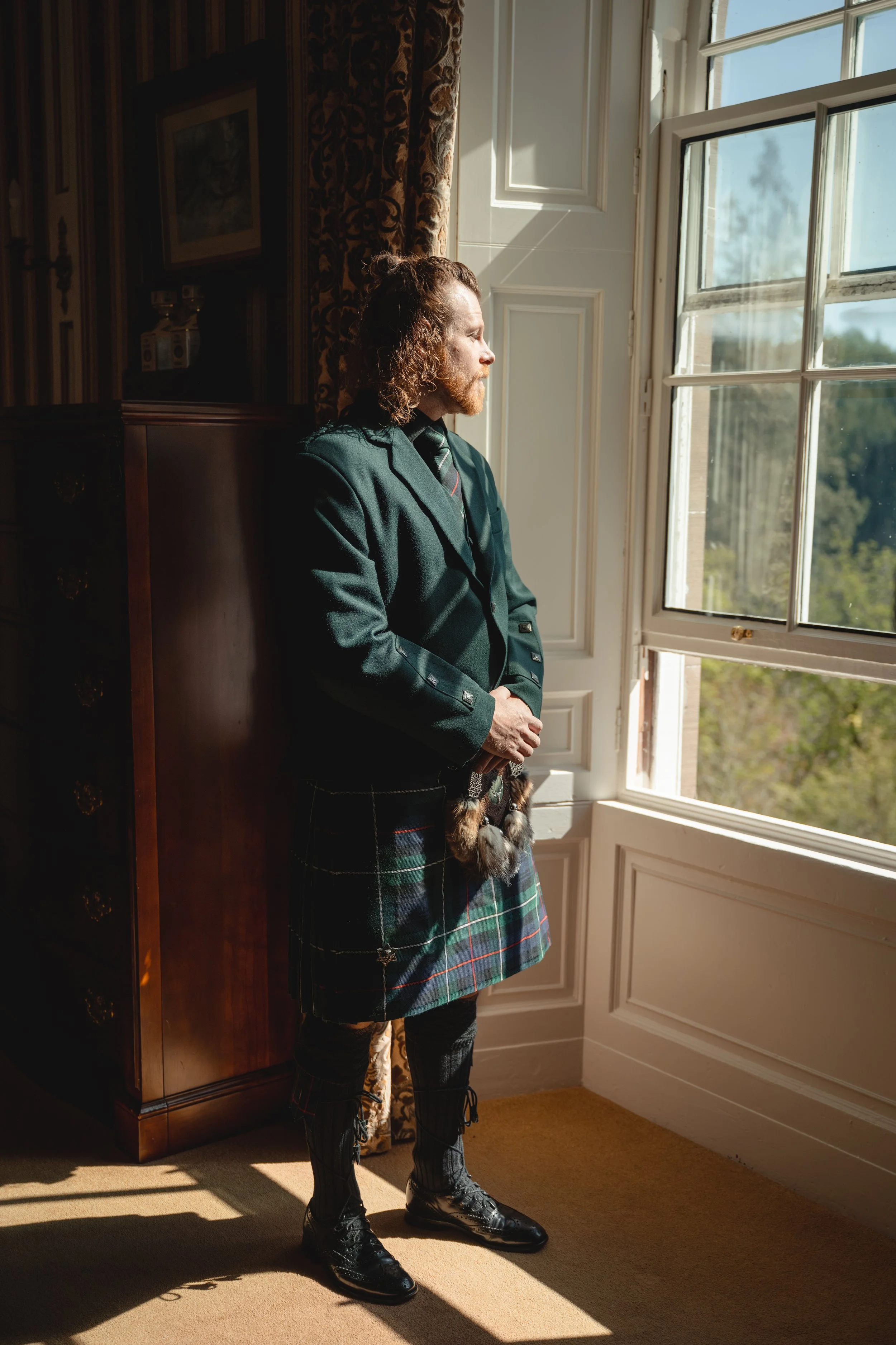 A man with curly hair and a beard stands indoors, looking out a window with sunlight on his face, wearing traditional Scottish attire including a kilt and a dark blazer.
