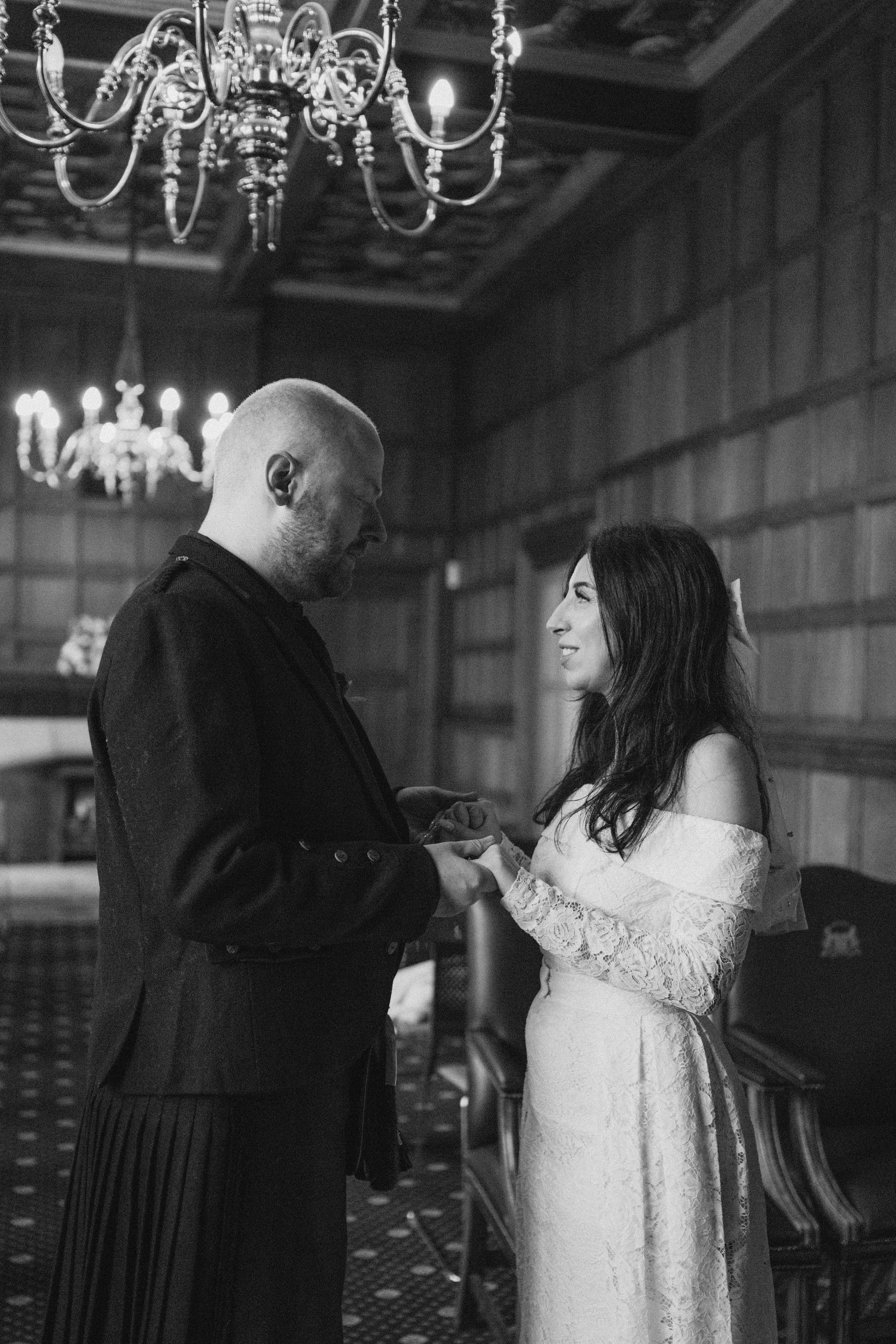 A black-and-white photo of a couple exchanging rings in an elegant, wood-paneled room with chandeliers hanging from the ceiling.
