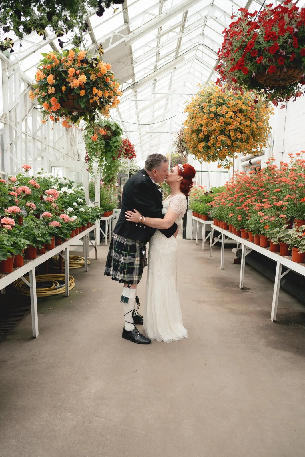 A bride and groom sharing a kiss in a greenhouse filled with hanging and potted flowers, including orange, pink, and red blooms.