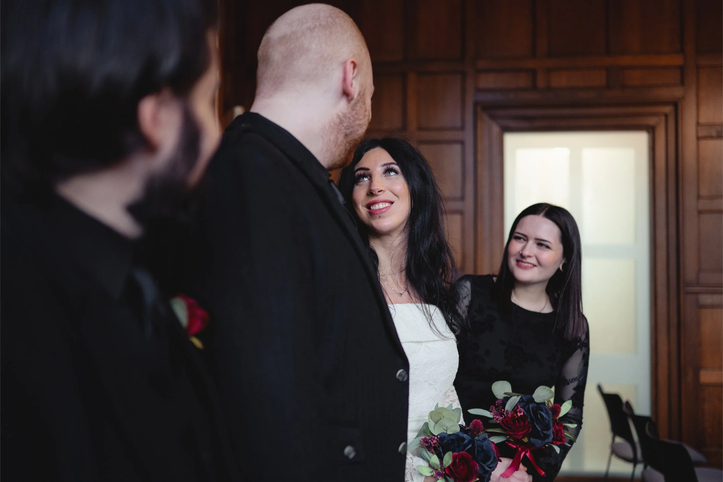 A woman in a white dress smiling up at a man with a bald head, dressed in black, during a wedding or formal event. Two other women, dressed in black, are standing behind her, one holding a bouquet of flowers and smiling.